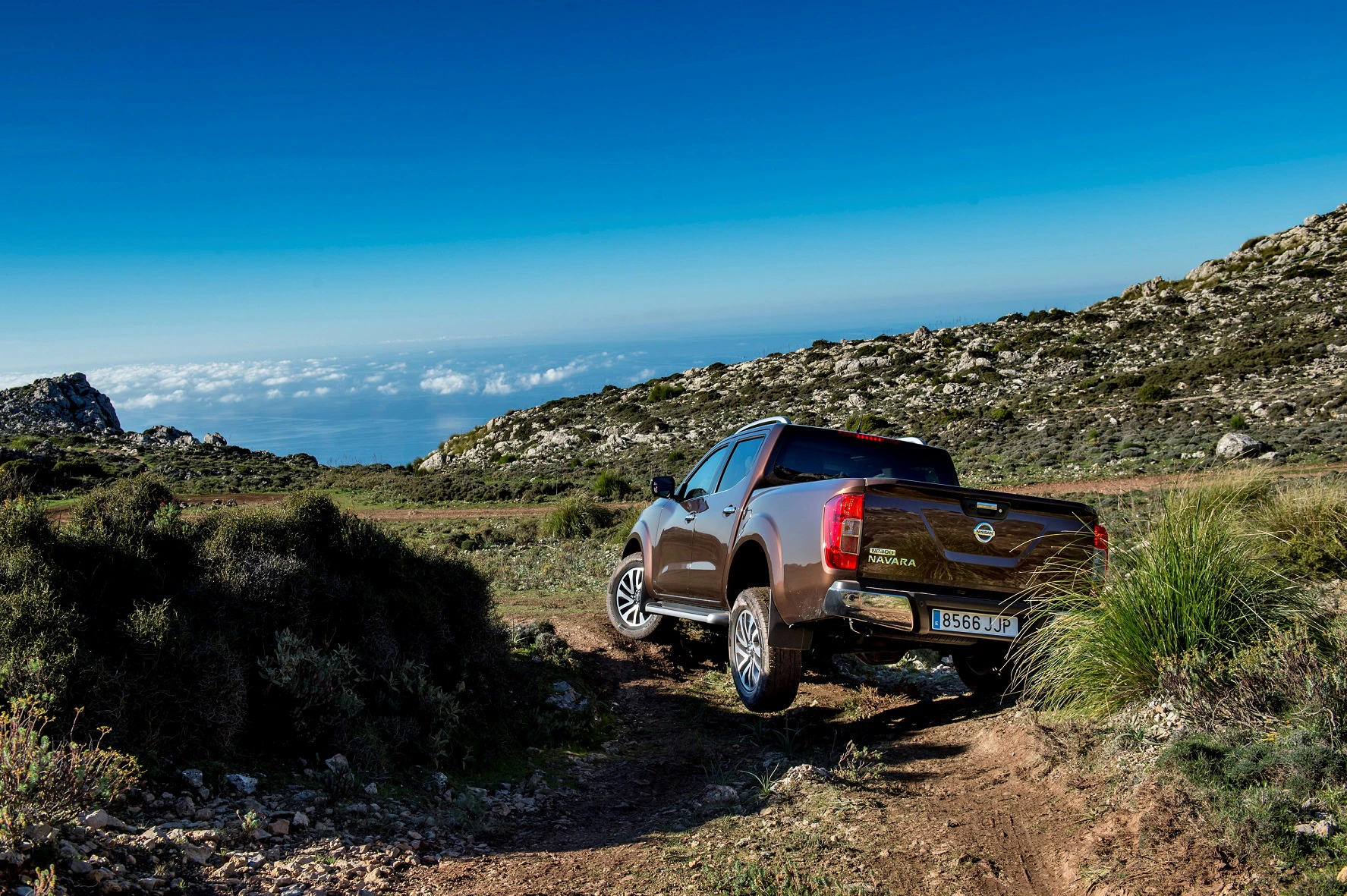 Ein brauner Pickup fährt auf einem steinigen Weg in einer bergigen Landschaft mit blauem Himmel und Blick auf das Meer im Hintergrund.