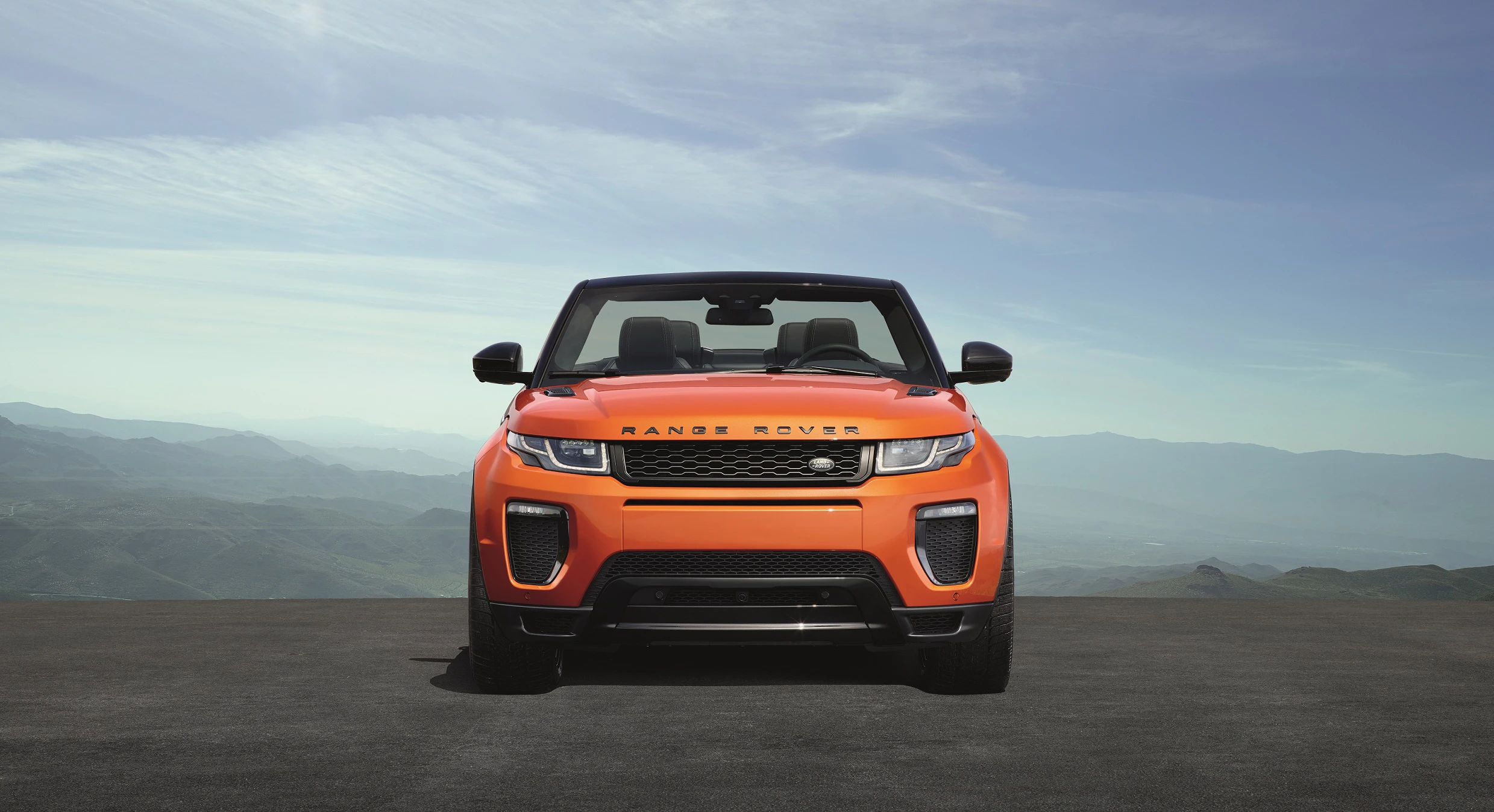 Orangefarbener Range Rover Cabriolet auf einer Straße vor einer Berglandschaft bei klarem Himmel.