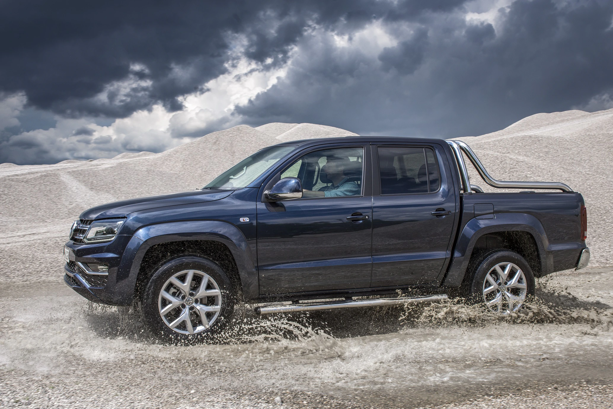 Ein blauer Pickup fährt durch Wasser auf einer Kiesstraße, umgeben von grauen Wolken und Schotterhügeln.