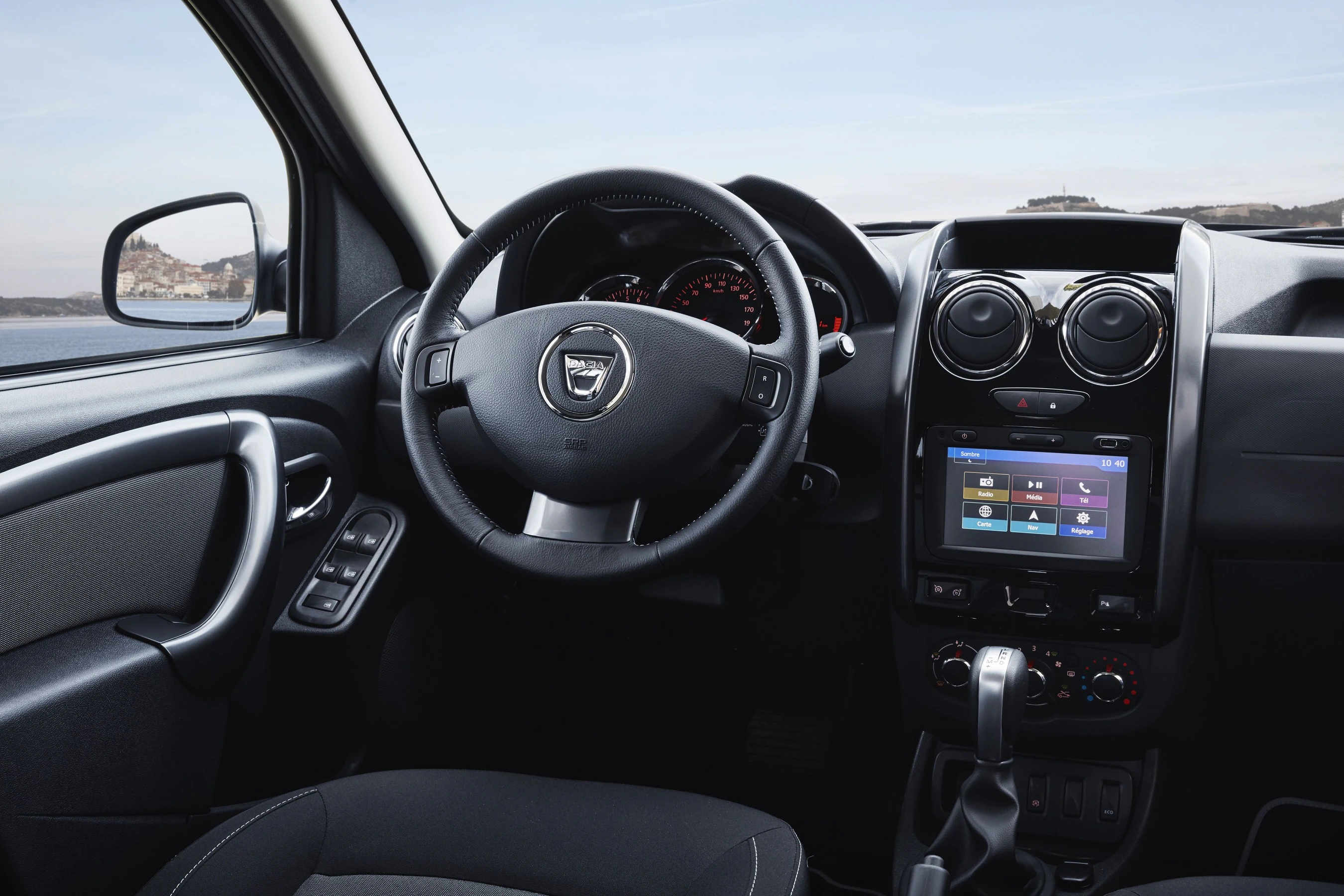 Innenraum eines Autos mit Lenkrad, Tachometer und zentralem Display mit Navigationsfunktionen. Außenlandschaft im Hintergrund sichtbar.