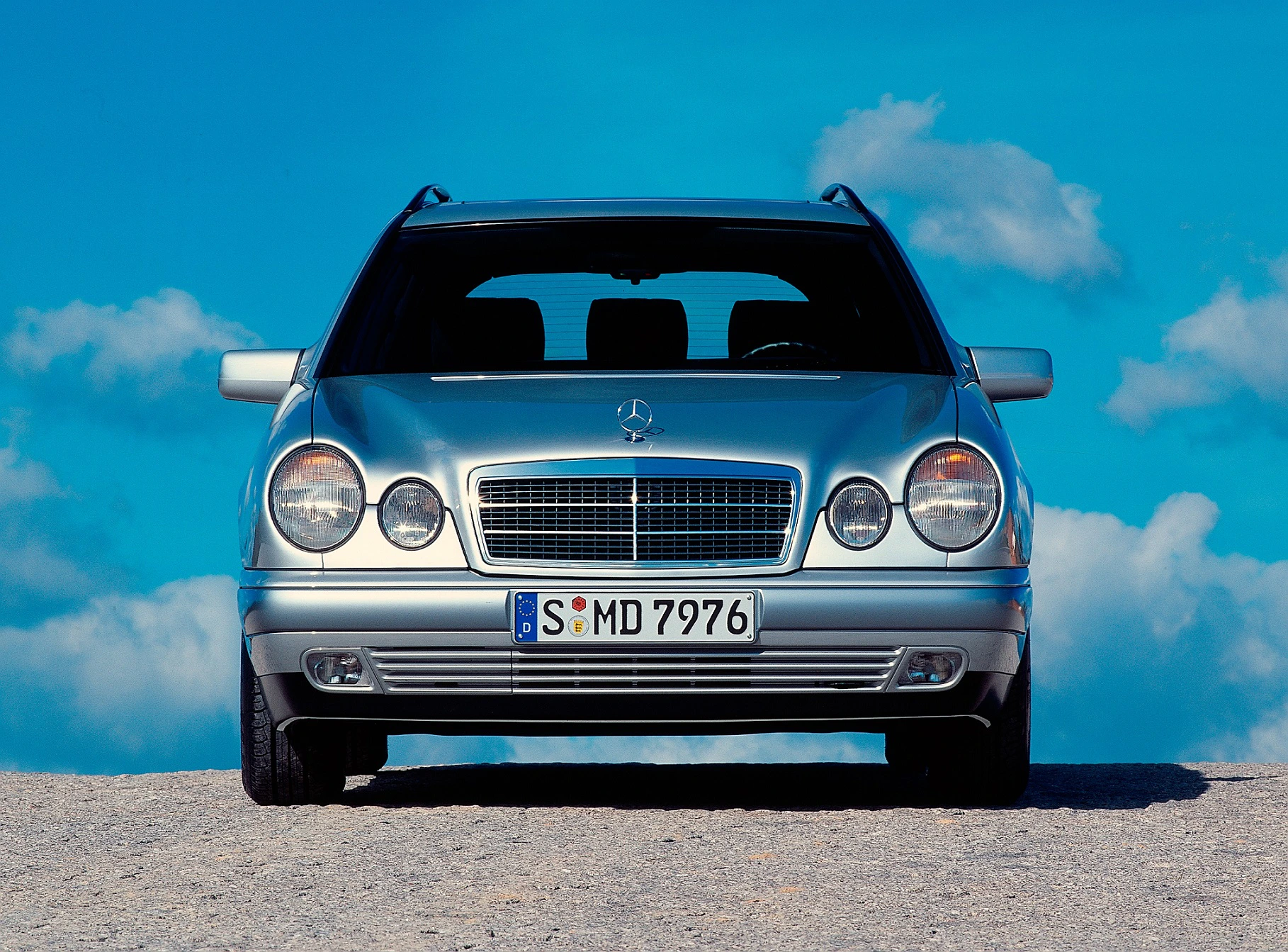 Silberner Mercedes-Benz-Kombi vor einem klaren blauen Himmel mit Wolken, frontal fotografiert.