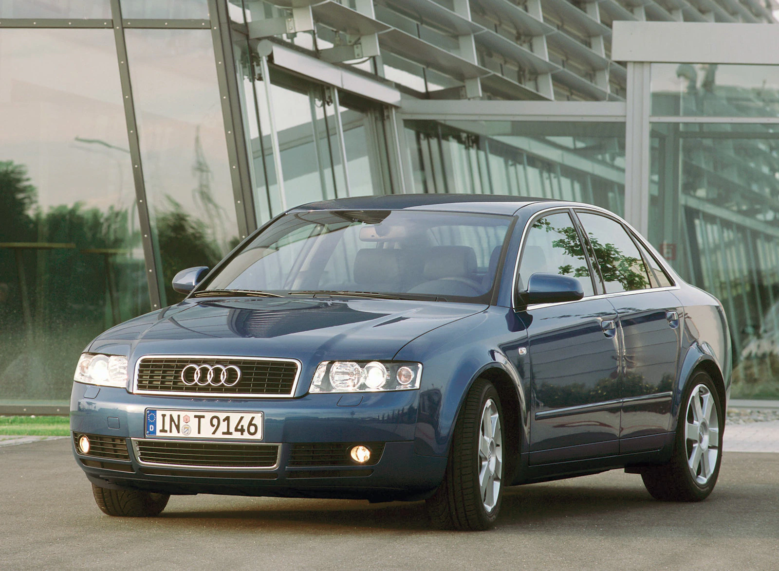 Dunkelblauer Audi A4 vor einem modernen Glasgebäude, seitliche Frontansicht, bei Tageslicht geparkt.