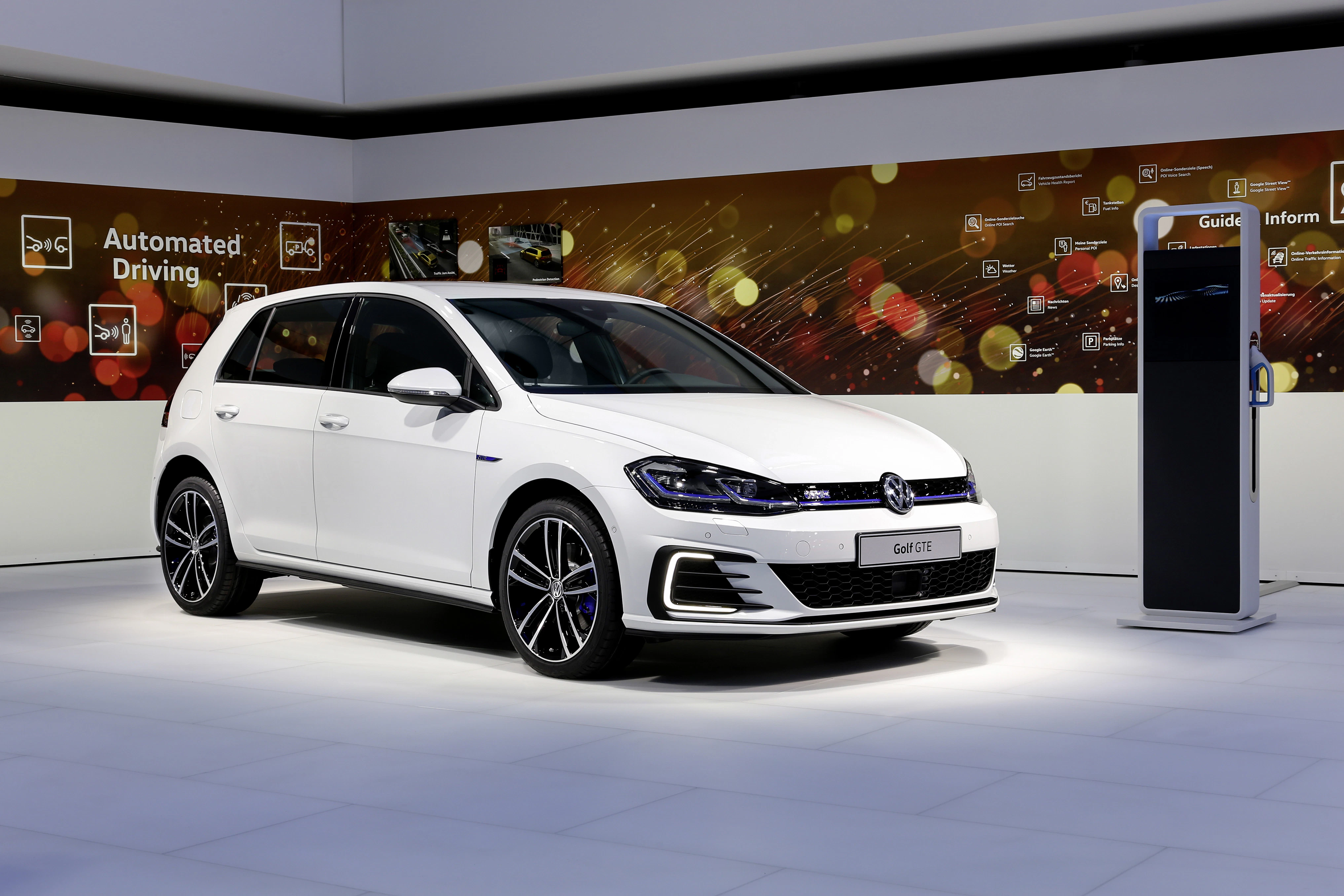 Weißer Volkswagen Golf auf einer Automobilausstellung, umgeben von modernen Grafiken und Lichtern.