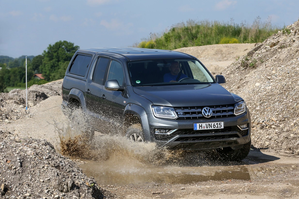 Dunkelgrauer VW-Pickup fährt durch Pfütze auf unbefestigtem Weg, spritzt Wasser in alle Richtungen. Hintergrund: hügelige Landschaft und blauer Himmel.
