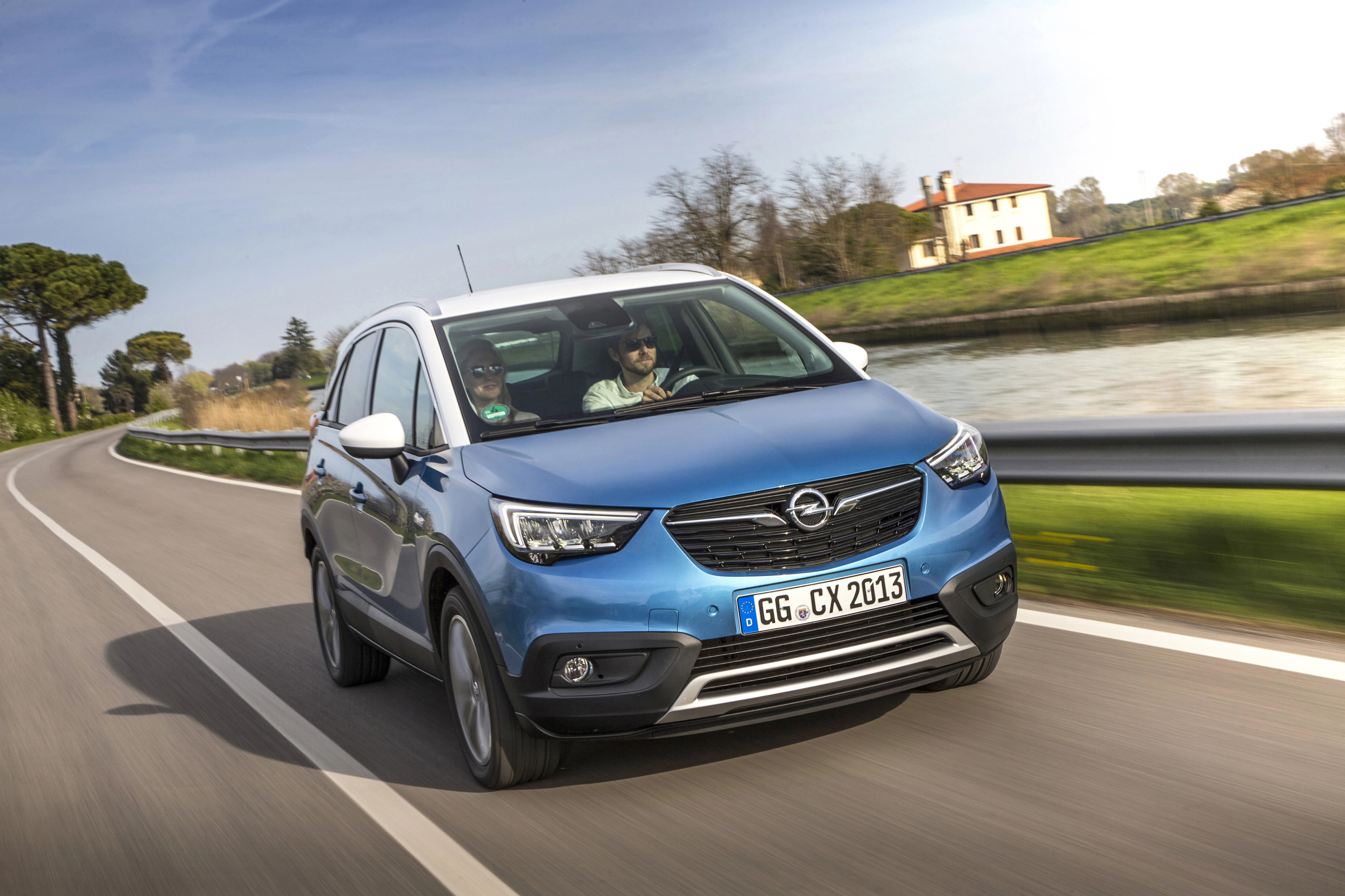 Blauer Opel Crossland X fährt auf einer Landstraße entlang eines Kanals, bei sonnigem Wetter und klarem Himmel.