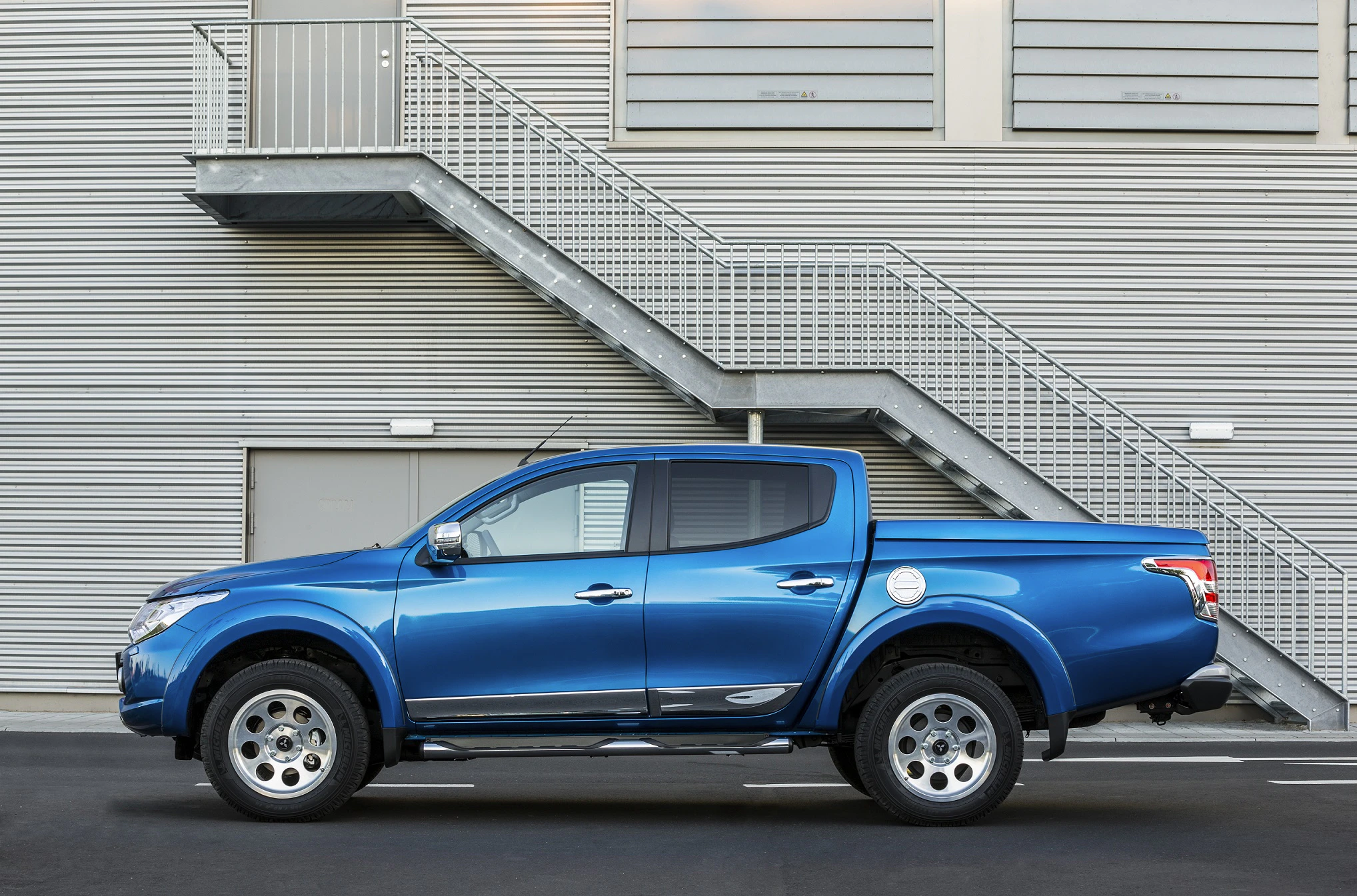 Blauer Pickup-Truck vor grauer Gebäude-Fassade mit Metalltreppe. Fahrzeug in Seitenansicht geparkt auf schwarzem Asphalt.