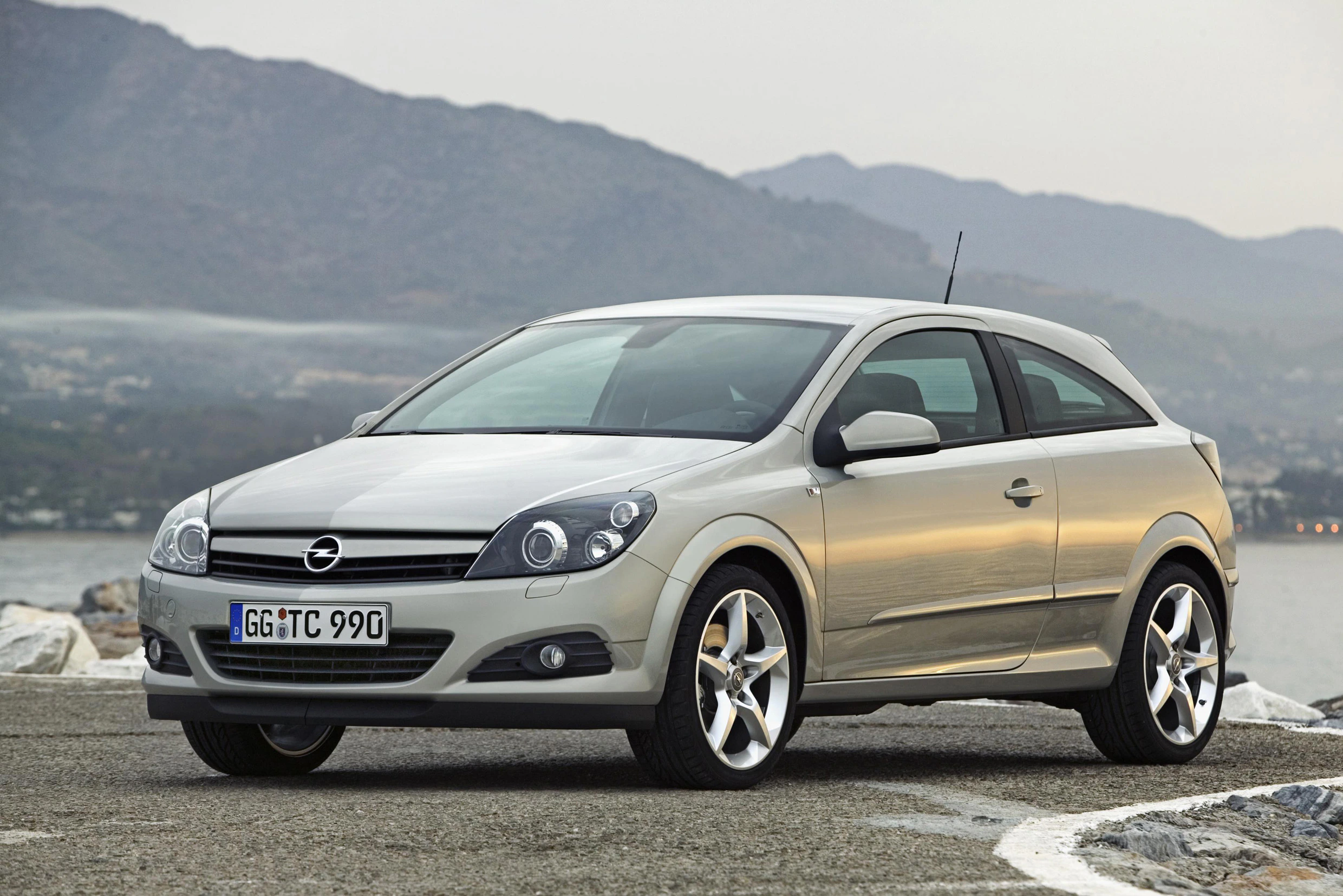 Silberner Opel Astra steht auf einer Küstenstraße vor bergiger Landschaft. Im Hintergrund ist das Meer zu sehen.