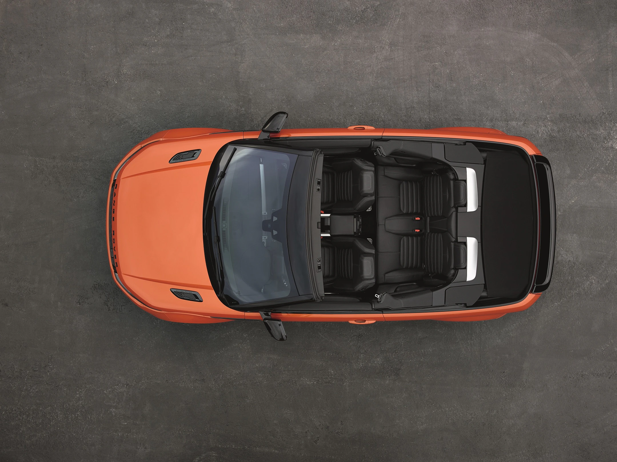 Orangenes Cabriolet von oben auf grauem Untergrund, Verdeck geöffnet, zeigt schwarze Ledersitze und Innenraumdetails.