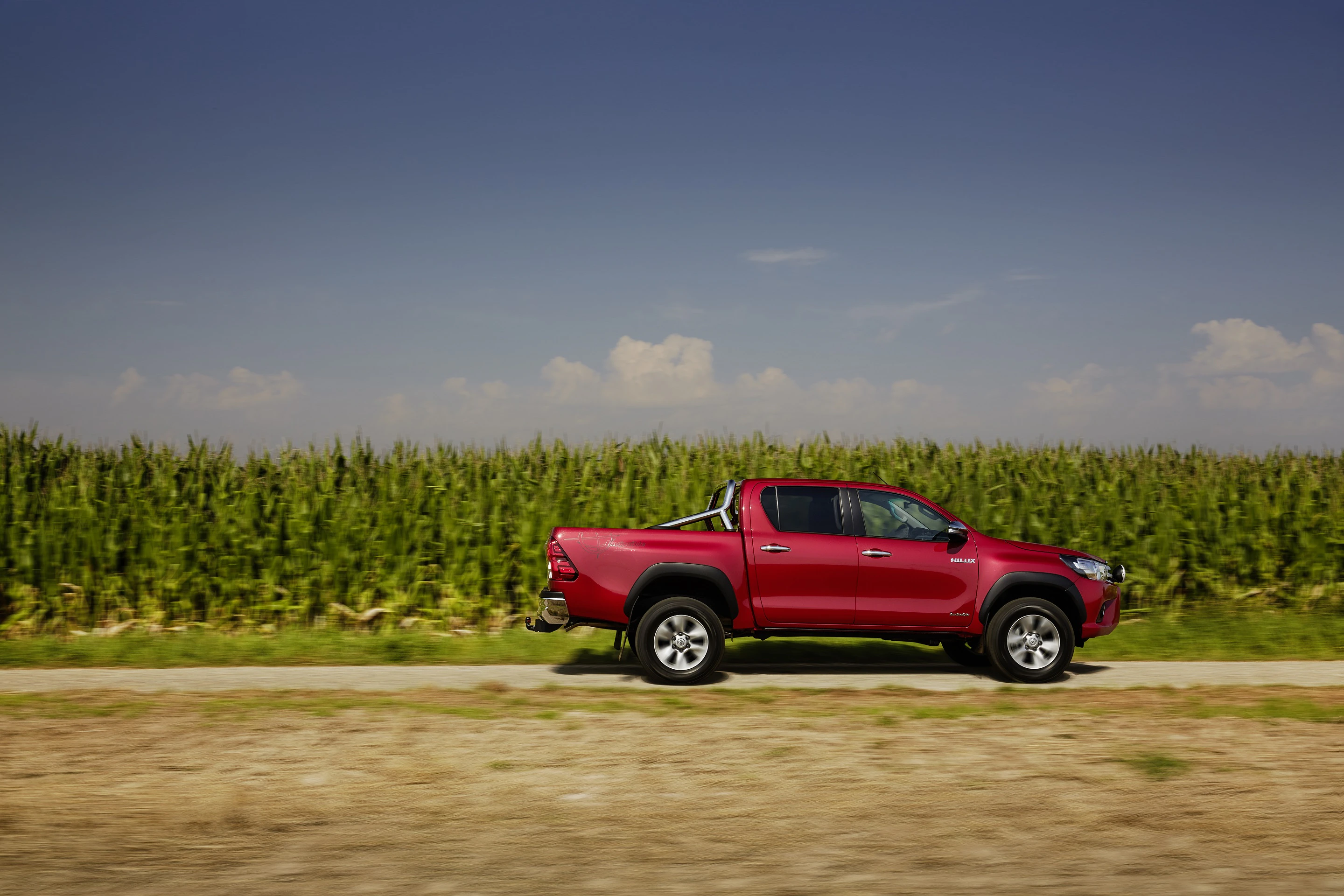 Roter Pickup fährt auf einer ländlichen Straße, im Hintergrund Maisfeld unter blauem Himmel.
