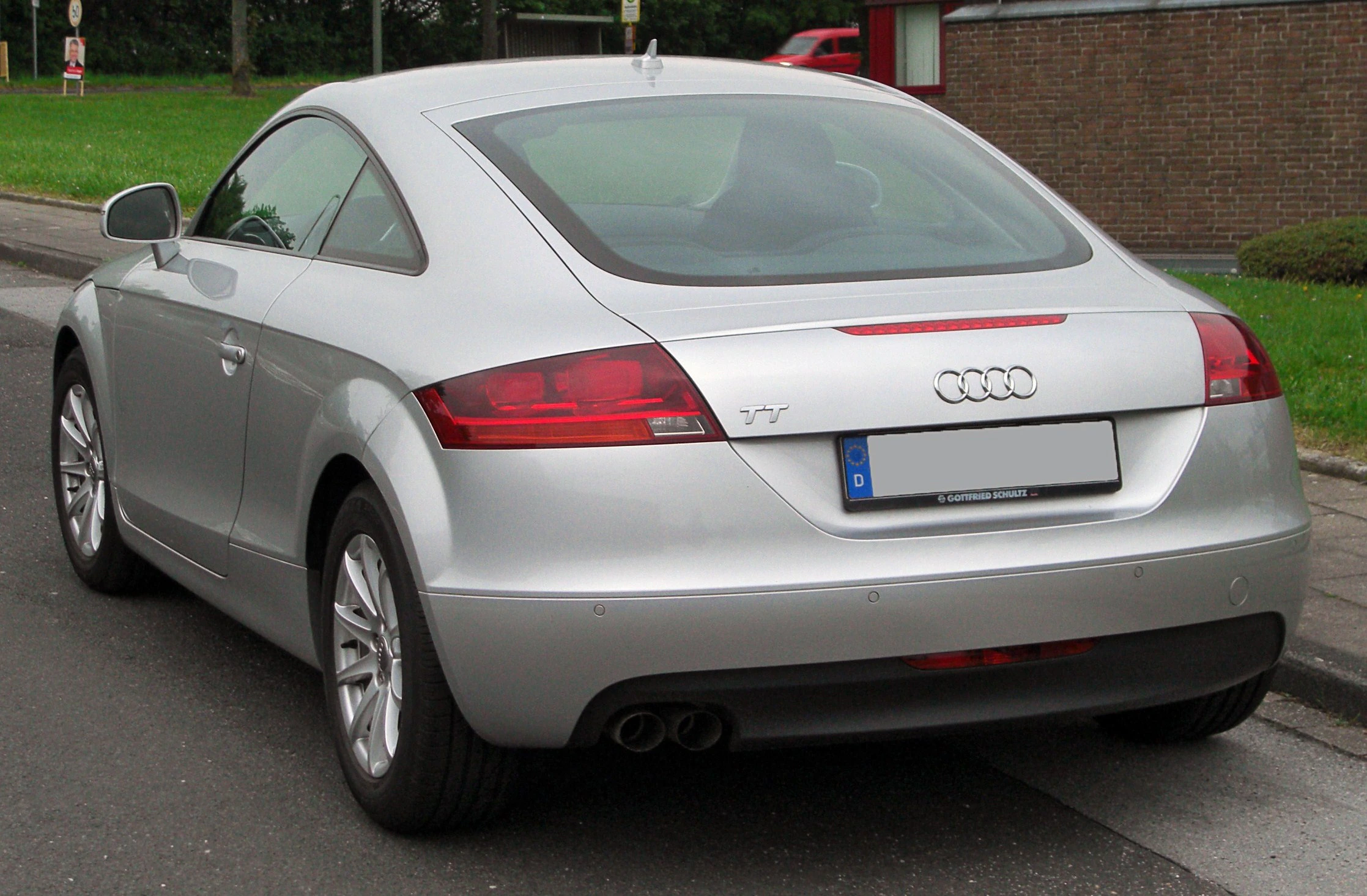 Silberner Audi TT, seitlich auf einer Straße geparkt, Hecktüransicht, mit sichtbaren Rücklichtern und Doppelauspuffen.