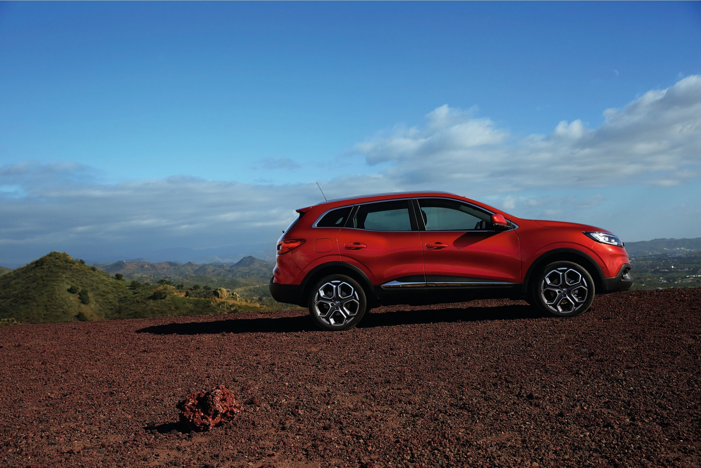 Roter SUV parkt auf einer Schotterstraße mit Blick auf bergige Landschaft und blauem Himmel im Hintergrund.