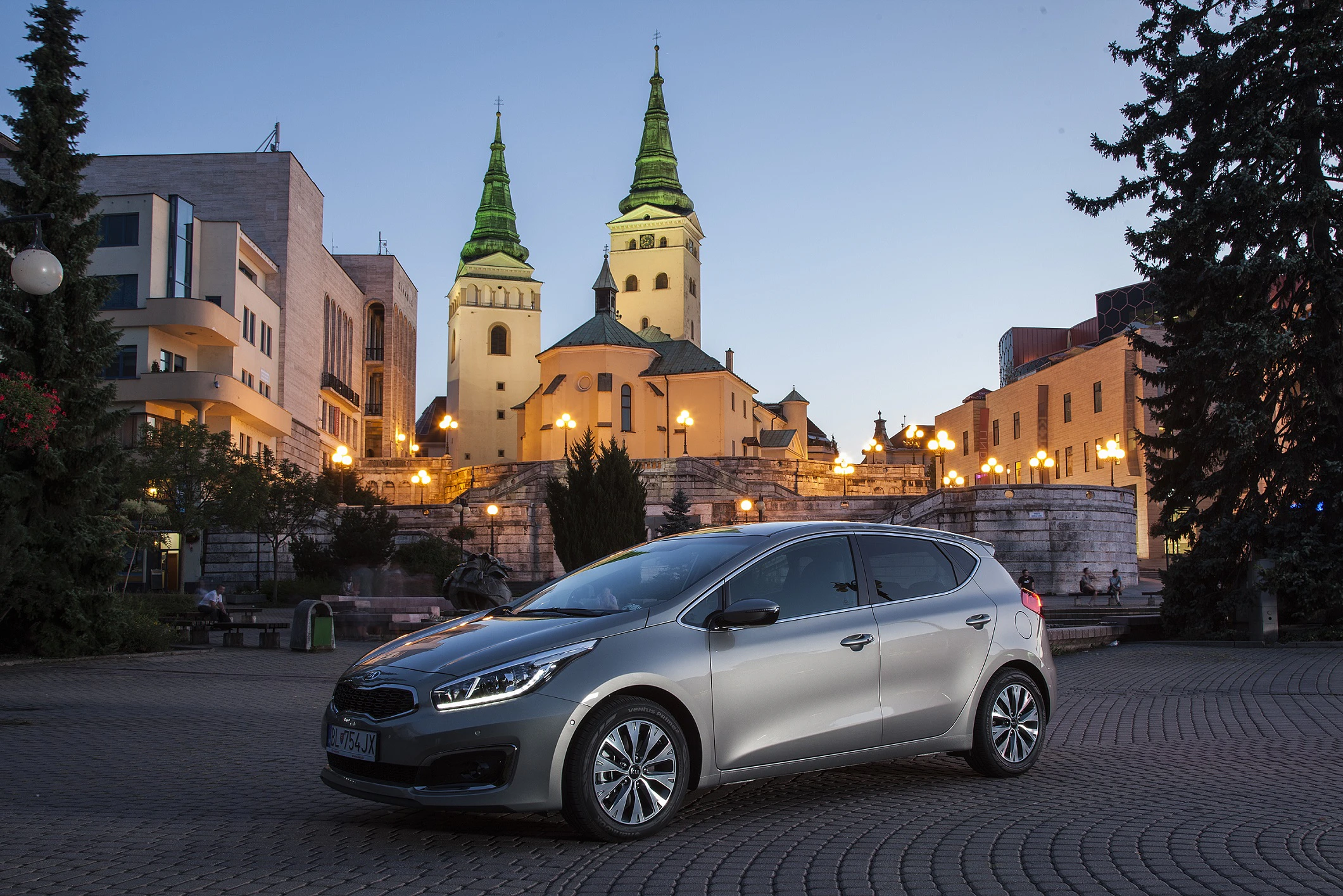 Silbernes Auto steht auf einem Platz bei Dämmerung; im Hintergrund beleuchtete Gebäude und eine Kirche mit zwei Türmen.