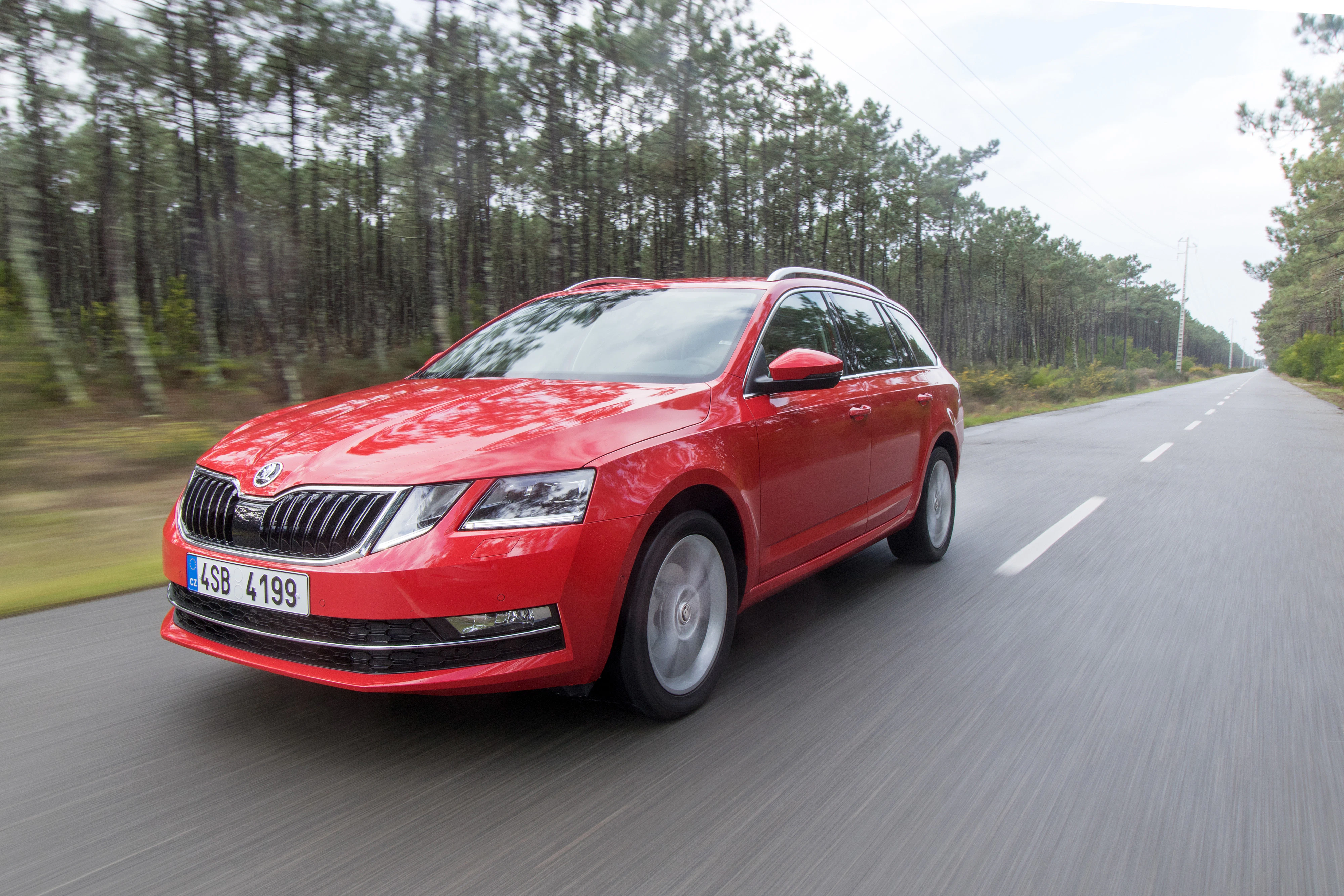 Roter Škoda Octavia Kombi fährt auf einer Landstraße, umgeben von Bäumen auf beiden Seiten. Die Straße ist leer und der Himmel bewölkt.