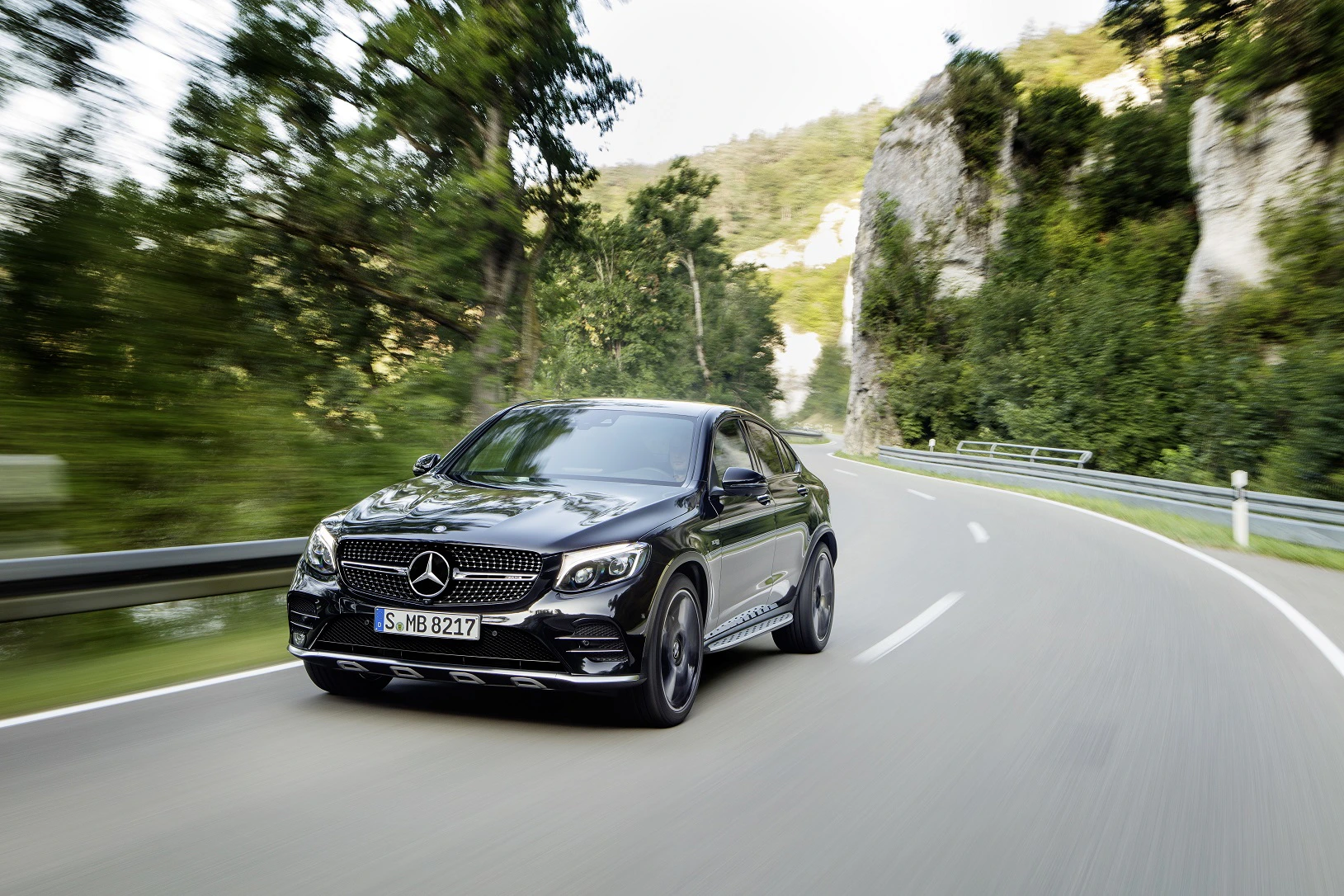 Schwarzes Mercedes-Benz Auto fährt schnell auf kurviger Landstraße, umgeben von Bäumen und Felsen bei Tageslicht.