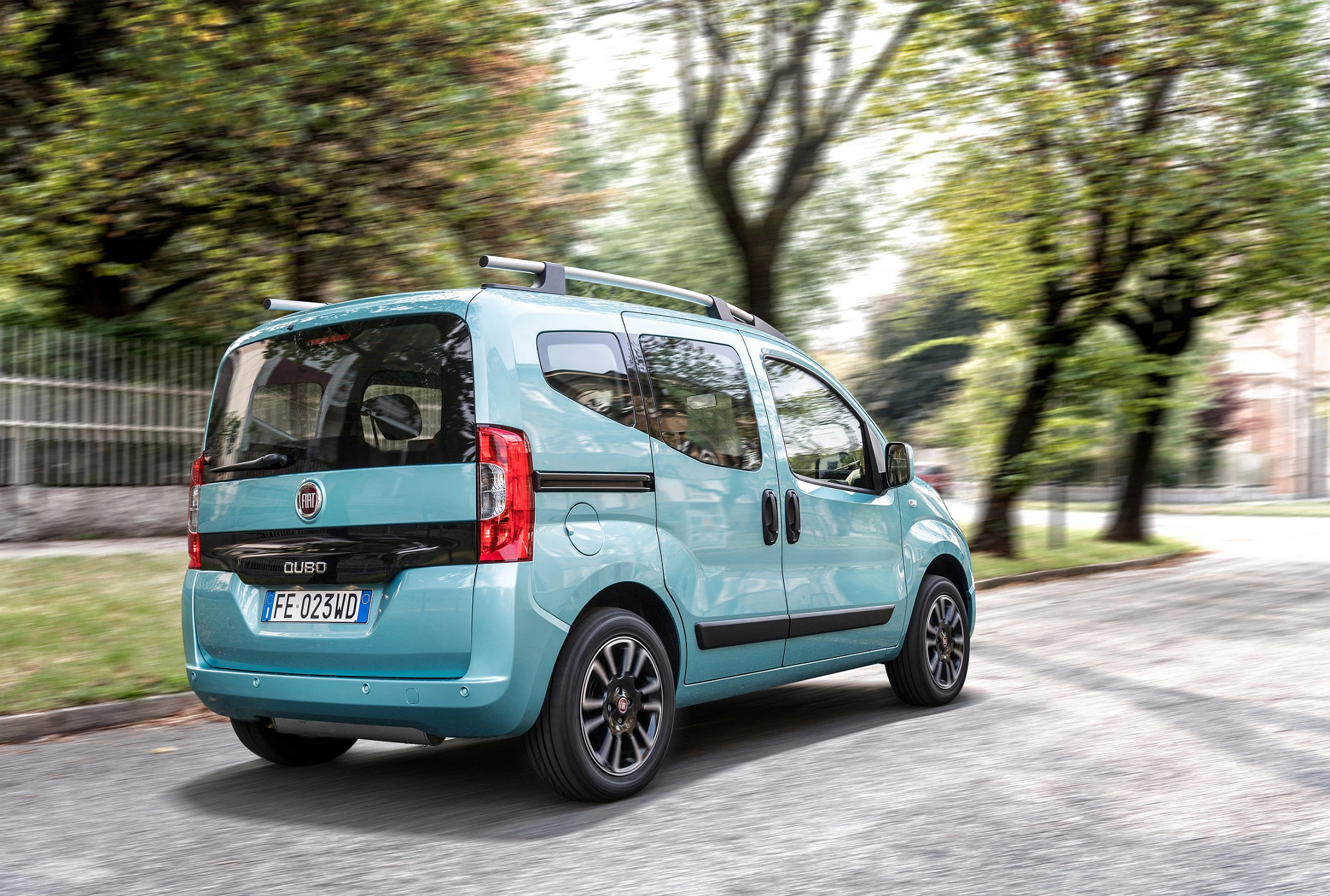 Ein hellblauer Fiat Qubo fährt auf einer von Bäumen gesäumten Straße. Der Himmel ist klar, und die Sonne scheint durch die Blätter.