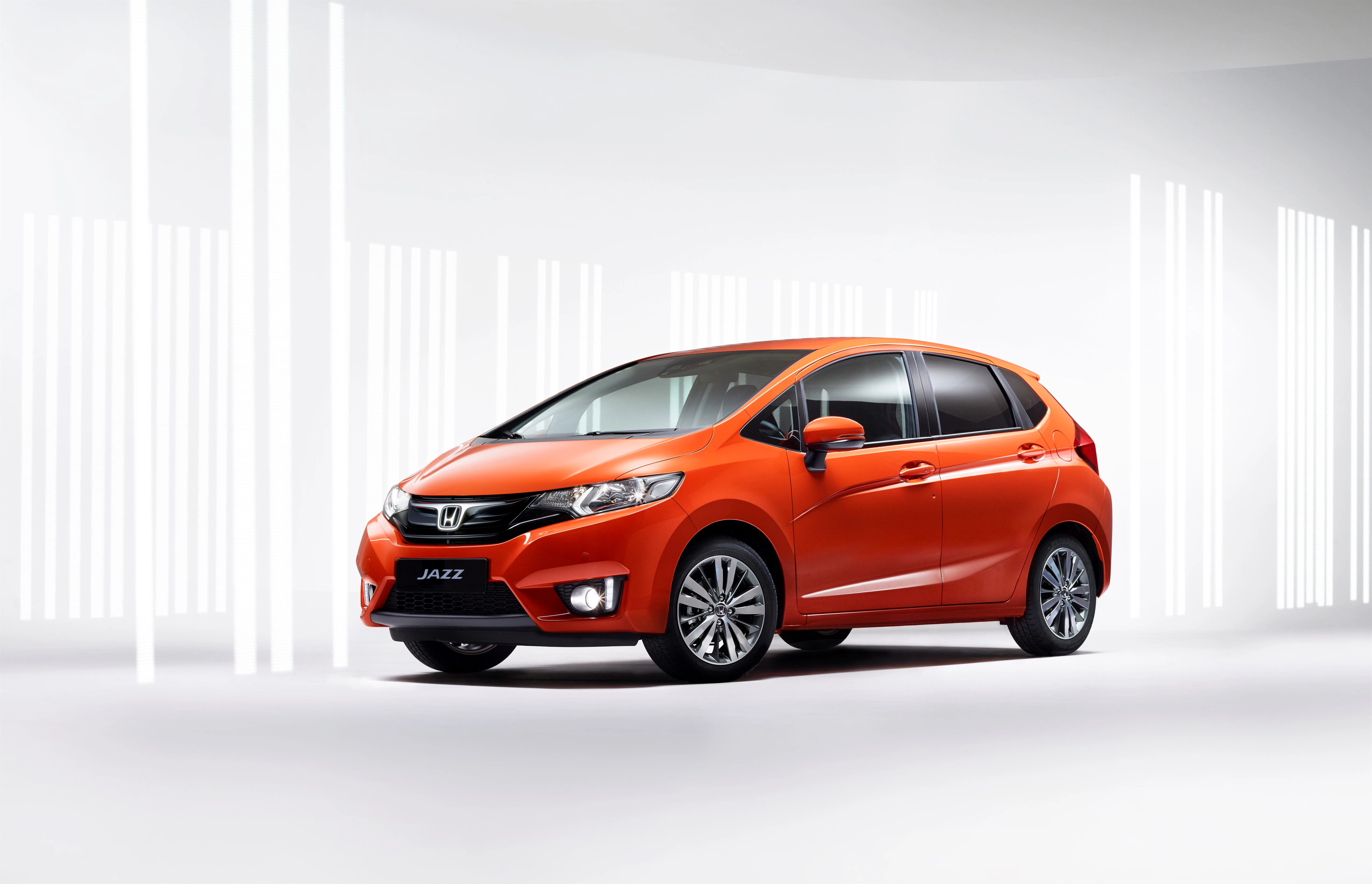 Oranger Honda Jazz in modernem, hell erleuchtetem Studio mit vertikalen Lichtstreifen im Hintergrund.