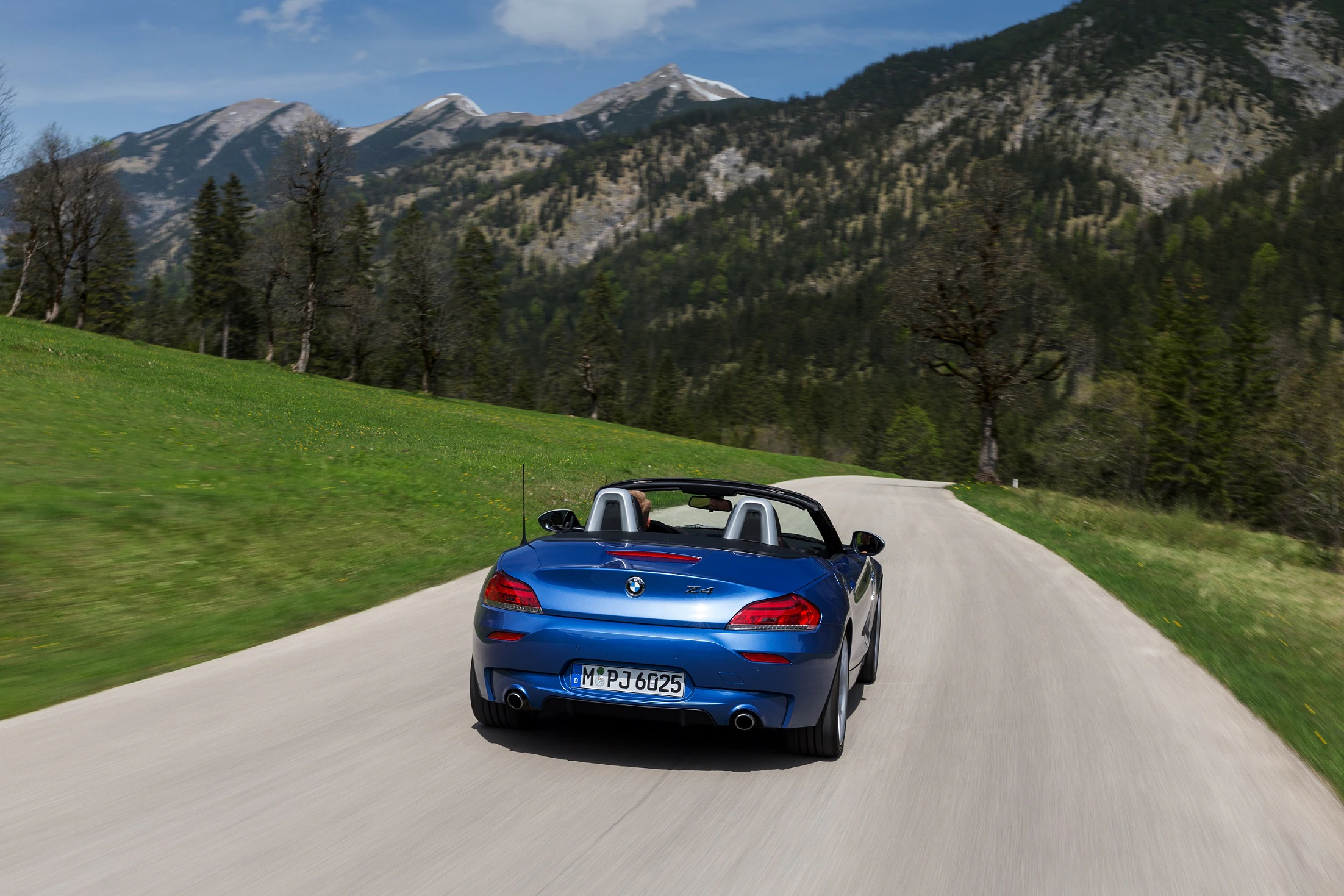 Blauer Cabrio-Sportwagen fährt auf einer kurvigen Straße durch malerische Berglandschaft mit grünen Wiesen und schneebedeckten Gipfeln im Hintergrund.