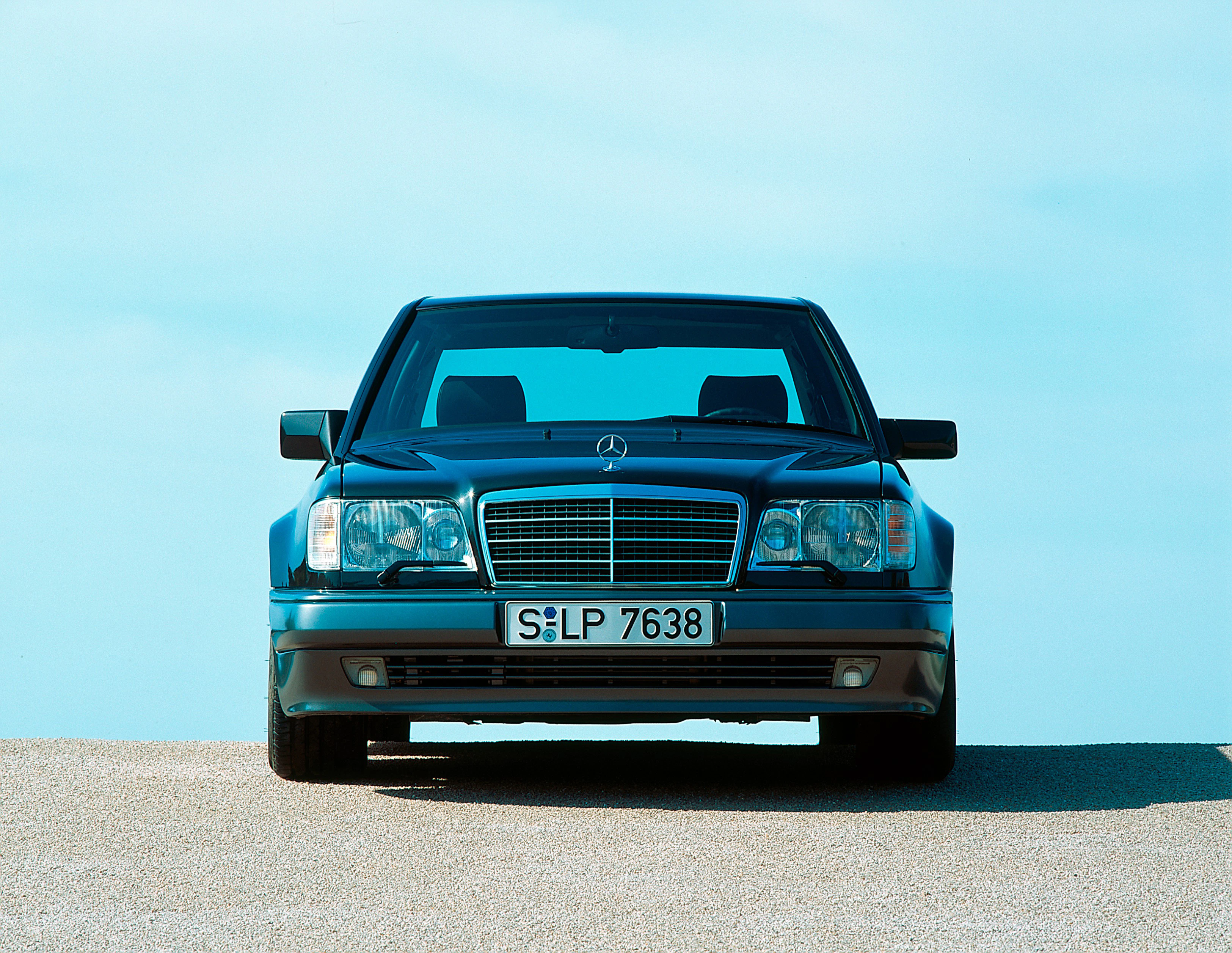Frontalansicht eines klassischen Mercedes-Benz W124 Limousine auf sandigem Boden vor einem blauen Himmel.