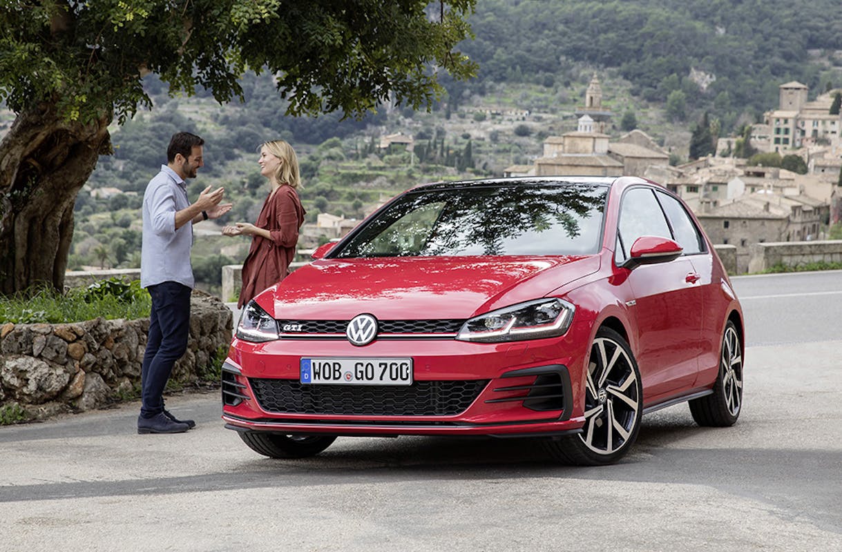 Ein roter VW Golf GTI steht auf einer Landstraße; im Hintergrund unterhalten sich zwei Personen vor einer bergigen Landschaft.