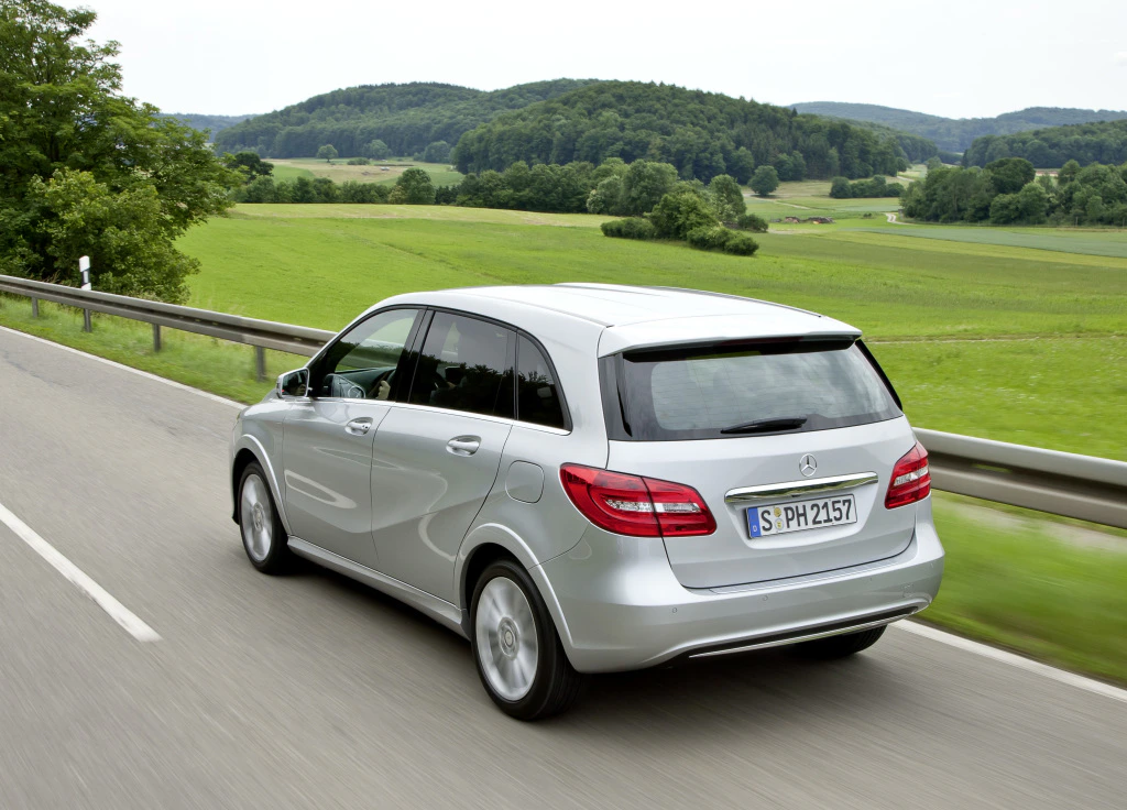 Silberner Mercedes-Benz fährt auf Landstraße in grüner, hügeliger Landschaft. Im Hintergrund sind Bäume und Wälder zu sehen.