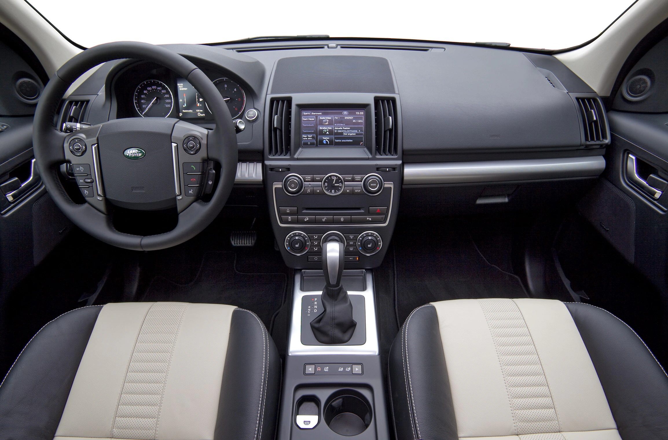 Innenraum eines Autos mit schwarzem und beigem Interieur, Lenkrad links, zentrale Steuerkonsole mit Bildschirm und Gangschaltung.