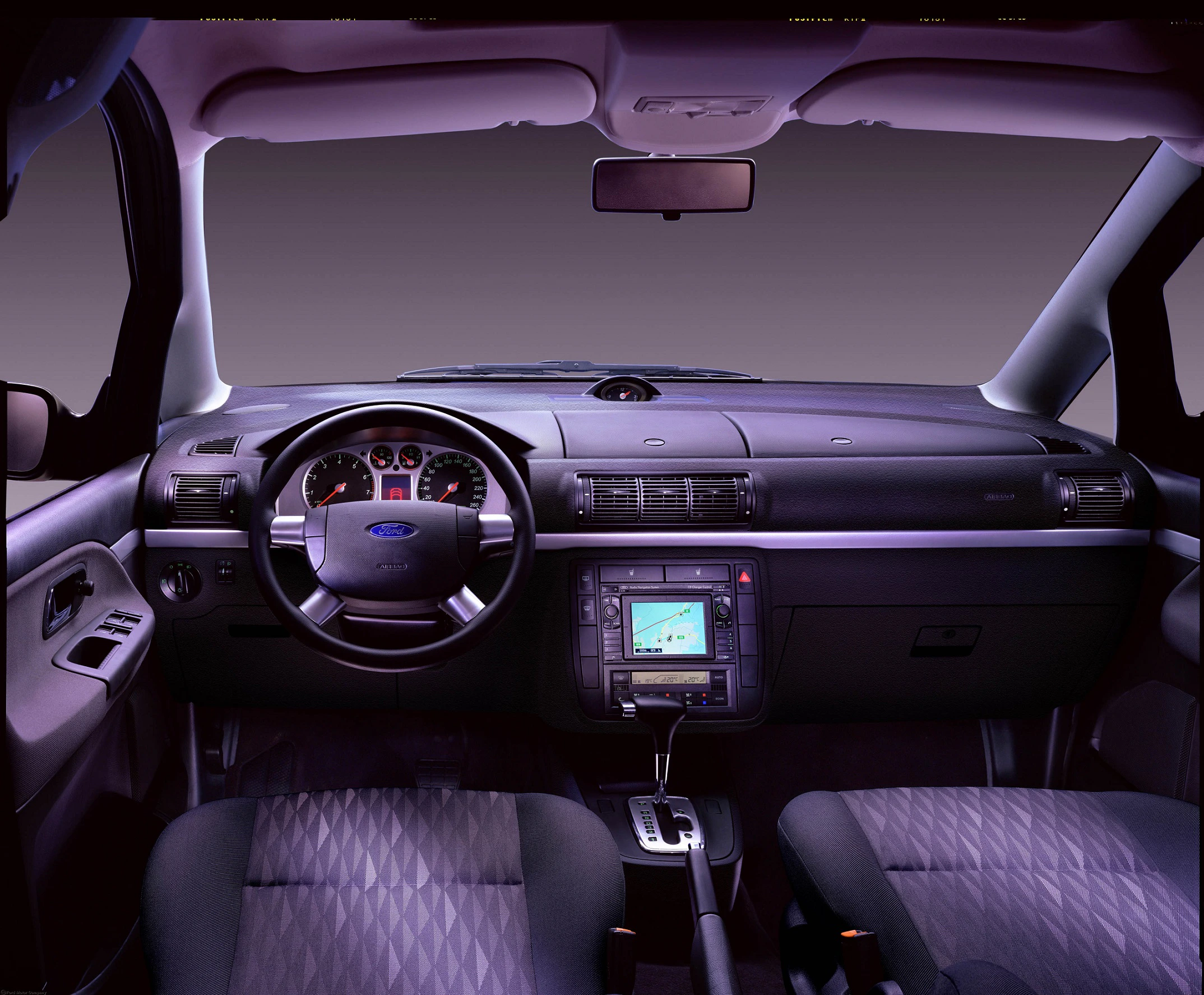 Innenraum eines Autos mit Lenkrad, Armaturenbrett und Navigationssystem. Dunkle Sitze und zentrale Konsole mit technischen Anzeigen.