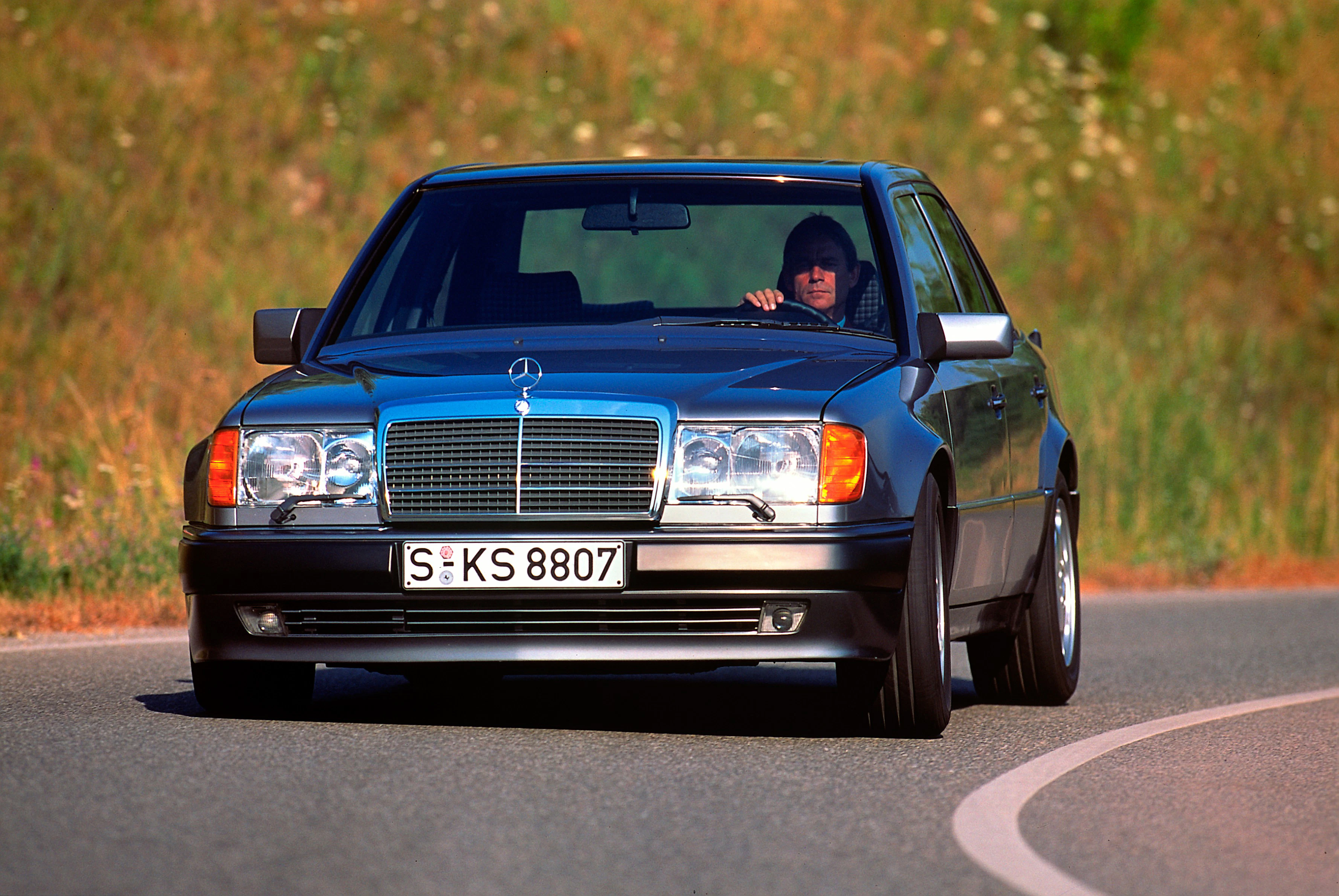 Schwarzer Mercedes-Benz W124 fährt auf einer kurvigen Landstraße. Im Hintergrund sind Gras und Wildblumen sichtbar. Fahrer blickt konzentriert.