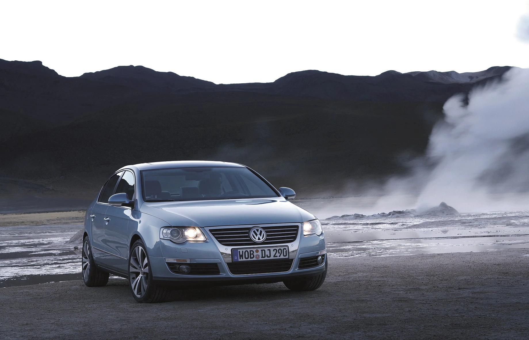 Silberner Volkswagen Passat geparkt vor einer dampfenden Landschaft mit Bergen im Hintergrund bei Sonnenaufgang.