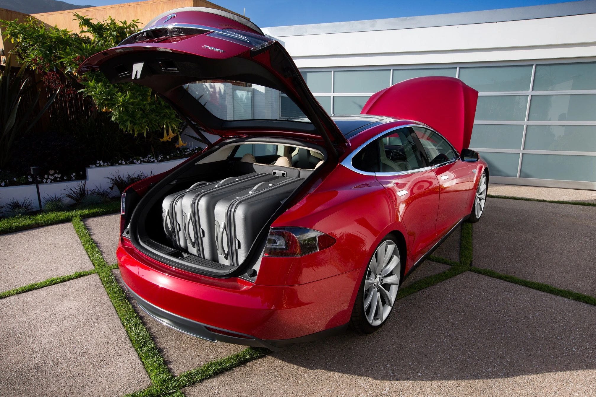 Ein rotes Auto mit geöffnetem Kofferraum und Frunk, beladen mit silbernen Koffern, steht auf einer betonierten Auffahrt.