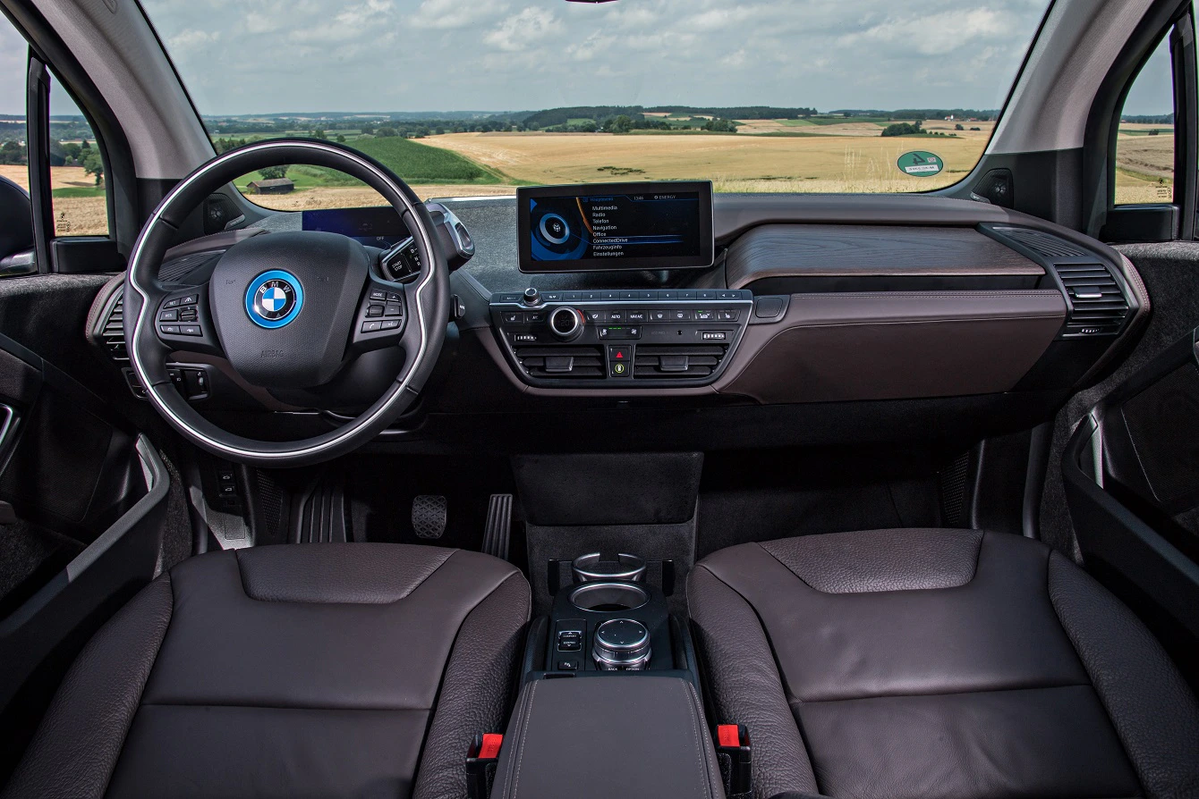 Innenraum eines BMW-Autos mit modernem Armaturenbrett, Lederlenkrad und integrierten Bildschirmen. Blick auf Felder durch die Windschutzscheibe.