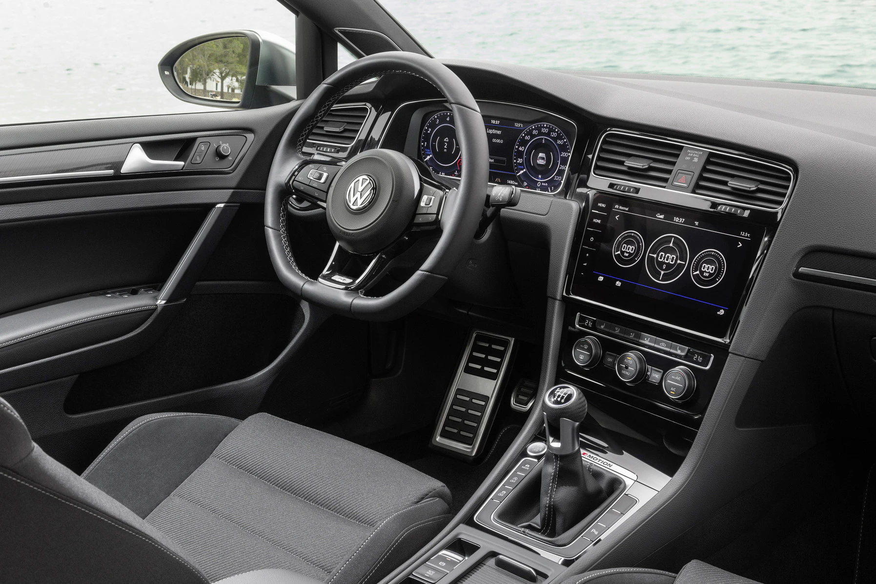 Innenraum eines modernen Volkswagen-Autos mit digitalem Cockpit, Lenkrad, Schalthebel und Infotainment-System, Blick auf See im Hintergrund.