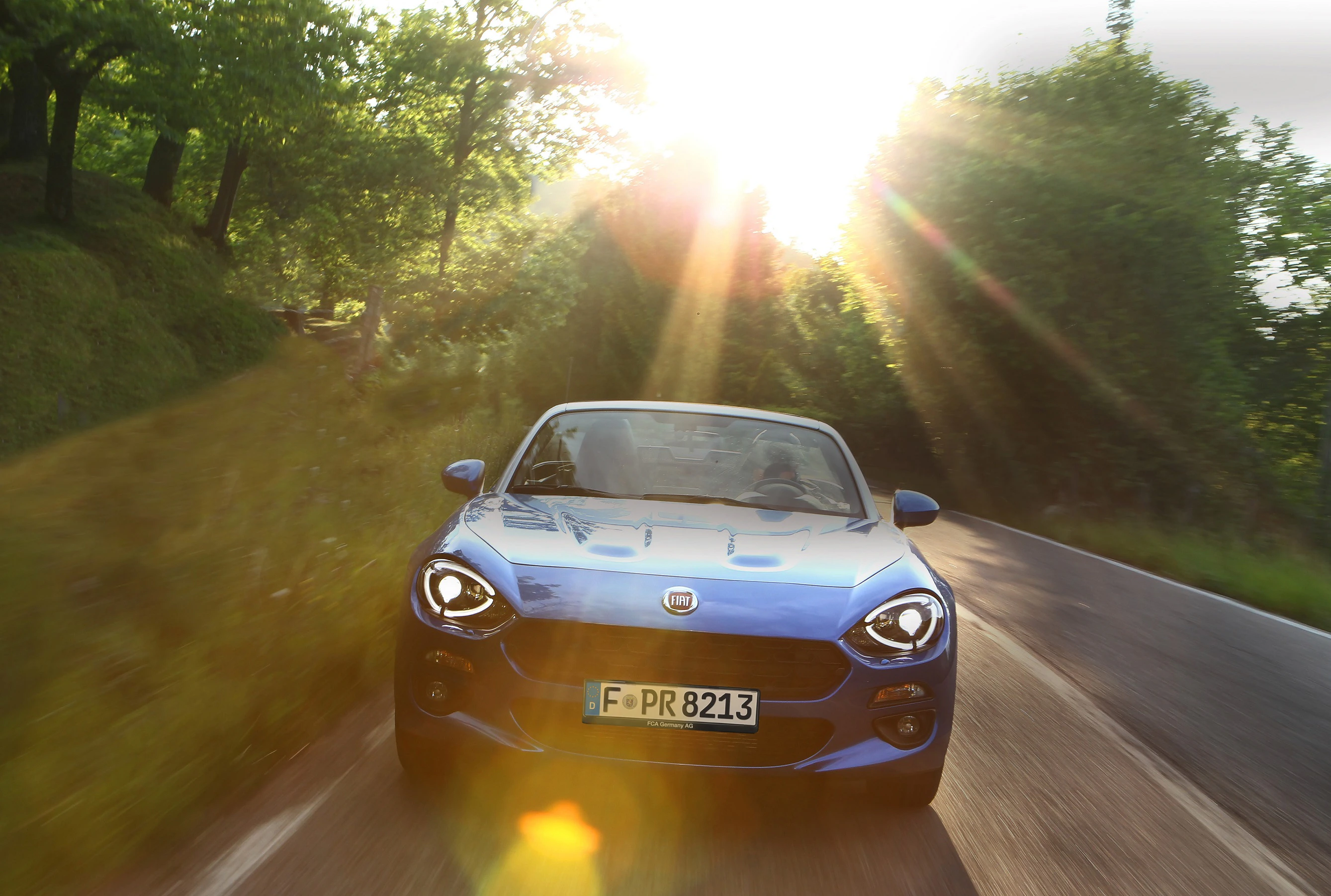 Ein blauer Fiat 124 Spider fährt bei Sonnenstrahlen auf einer kurvigen Landstraße, umgeben von grünen Bäumen.