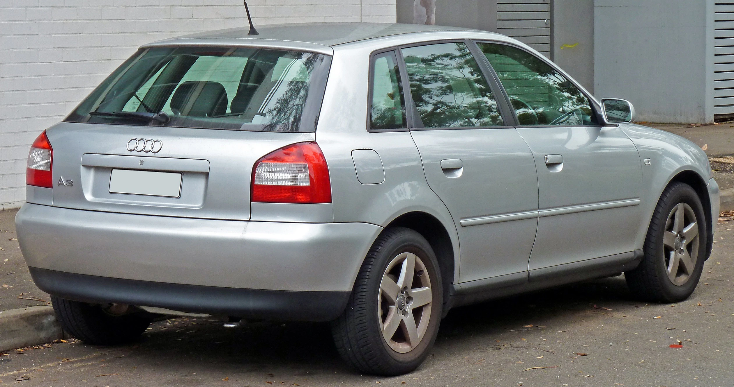 Silberner Audi A3, geparkt auf einer Straße, Seiten- und Heckansicht, mit mehrstöckigen Gebäude im Hintergrund.