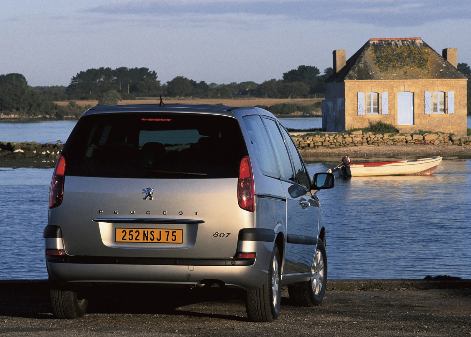 Silberner Peugeot 807 steht an einem Gewässer mit kleinem Steinhaus und Boot im Hintergrund. Ruhige, idyllische Szenerie.
