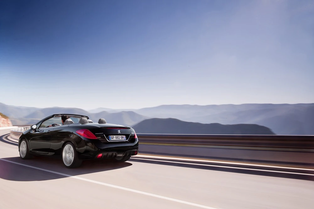 Schwarzer Cabrio-Sportwagen fährt auf kurvenreicher Bergstraße; blauer Himmel und verschwommene Gebirgslandschaft im Hintergrund.