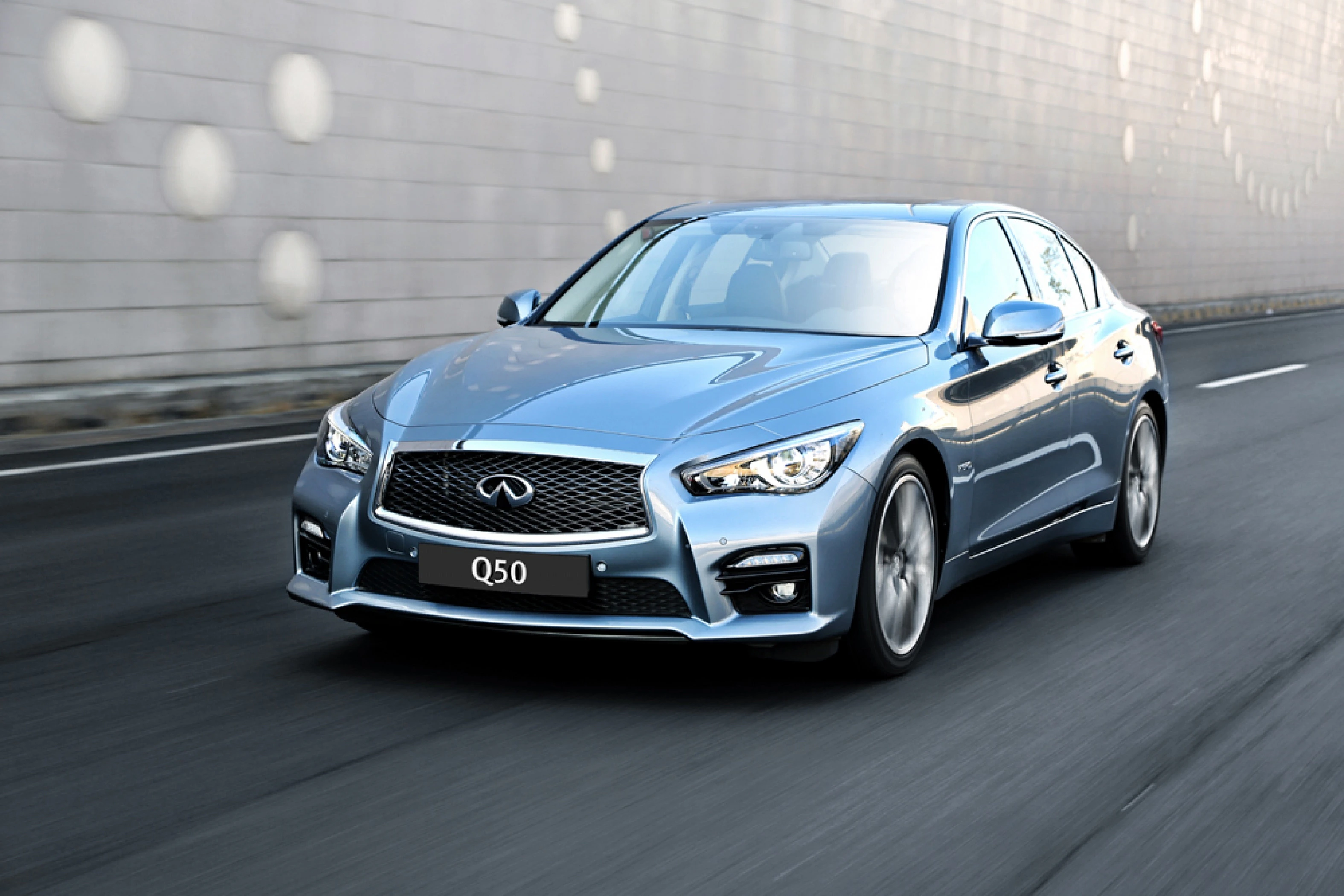 Blauer Infiniti Q50 fährt auf einer Straße vor einer gemusterten Wand. Modernes Design, Scheinwerfer eingeschaltet, dynamische Bewegung.