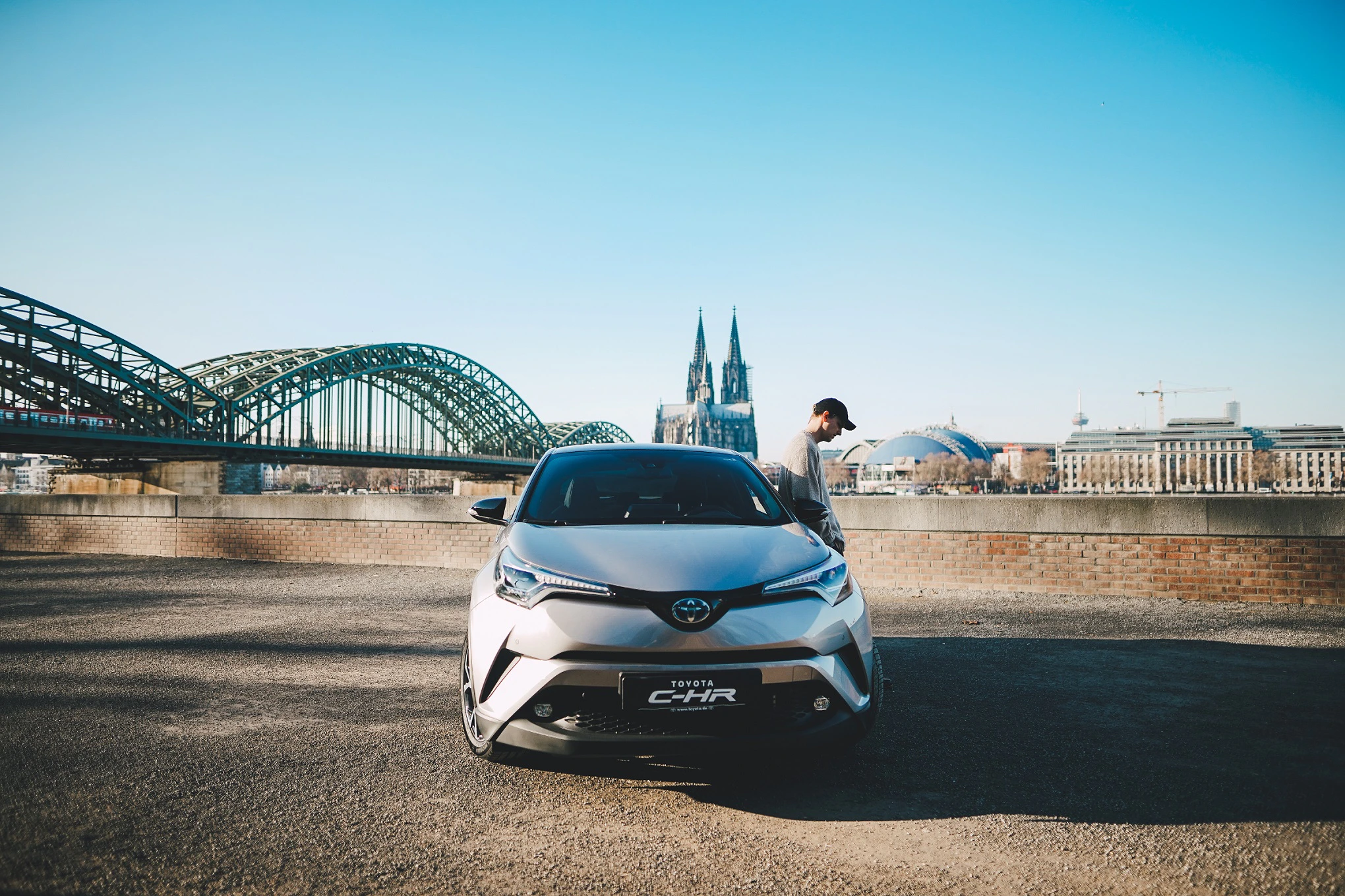 Silberner Toyota C-HR vor Kölner Dom und Hohenzollernbrücke, blauer Himmel, Person steht daneben.