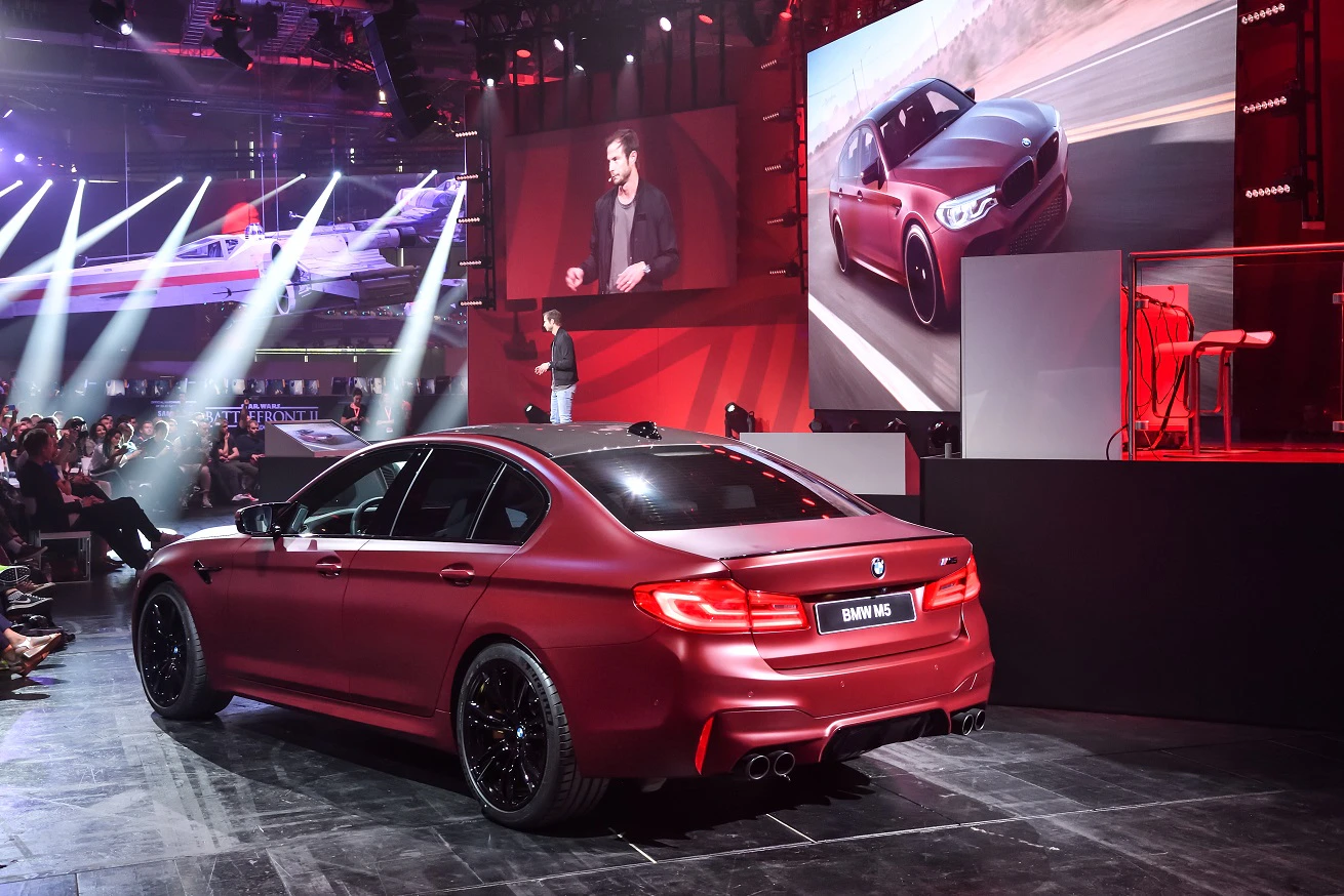 Roter BMW M5 auf einer Bühne, umgeben von Publikum und roten Lichtern. Präsentation mit Leinwandbild im Hintergrund.