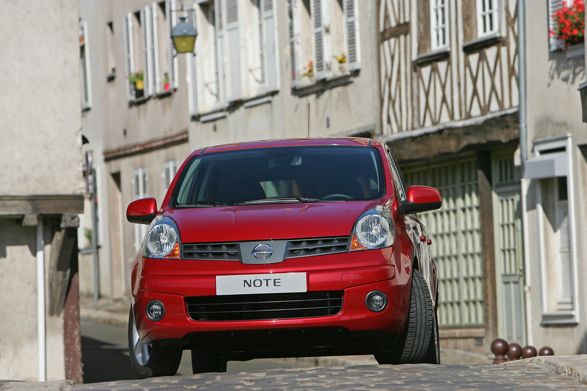 Rotes Auto, Nissan Note, auf gepflasterter Straße in malerischem, historischem Dorf mit Fachwerkhäusern und Blumen in den Fenstern.