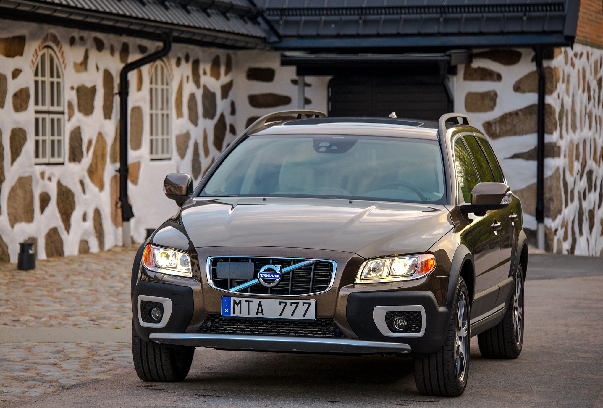Brauner Volvo XC70 steht vor einem steinverkleideten Gebäude mit schwarzen Fensterrahmen und Dach.