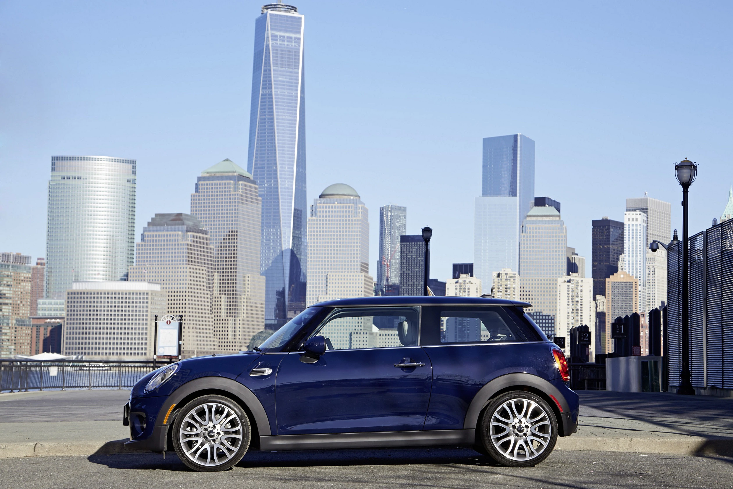 Blauer Mini Cooper steht vor der beeindruckenden Skyline von New York City mit modernen Wolkenkratzern im Hintergrund.