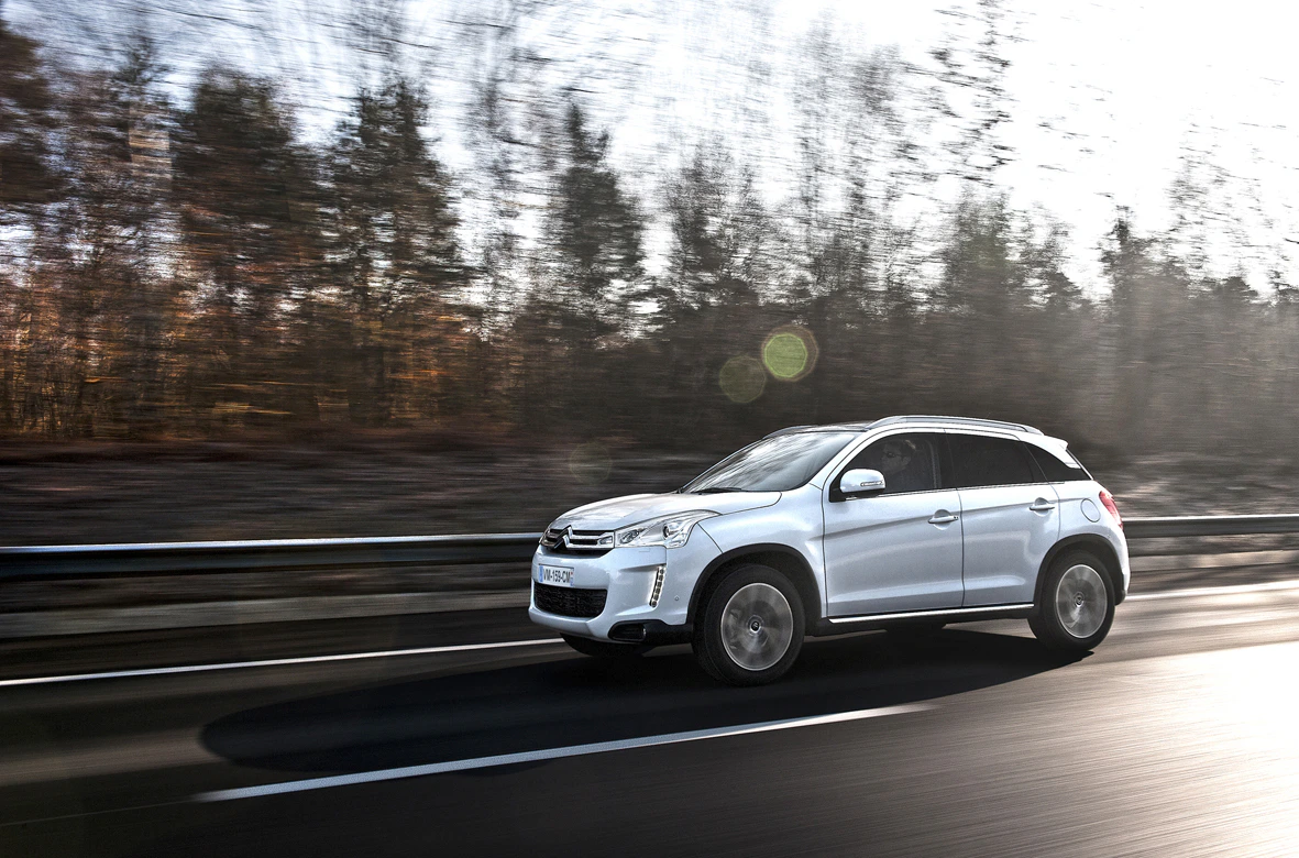 Silberner SUV fährt auf einer Landstraße entlang eines Waldes bei Sonnenschein. Im Hintergrund sind unscharfe Bäume und Lichtreflexionen sichtbar.