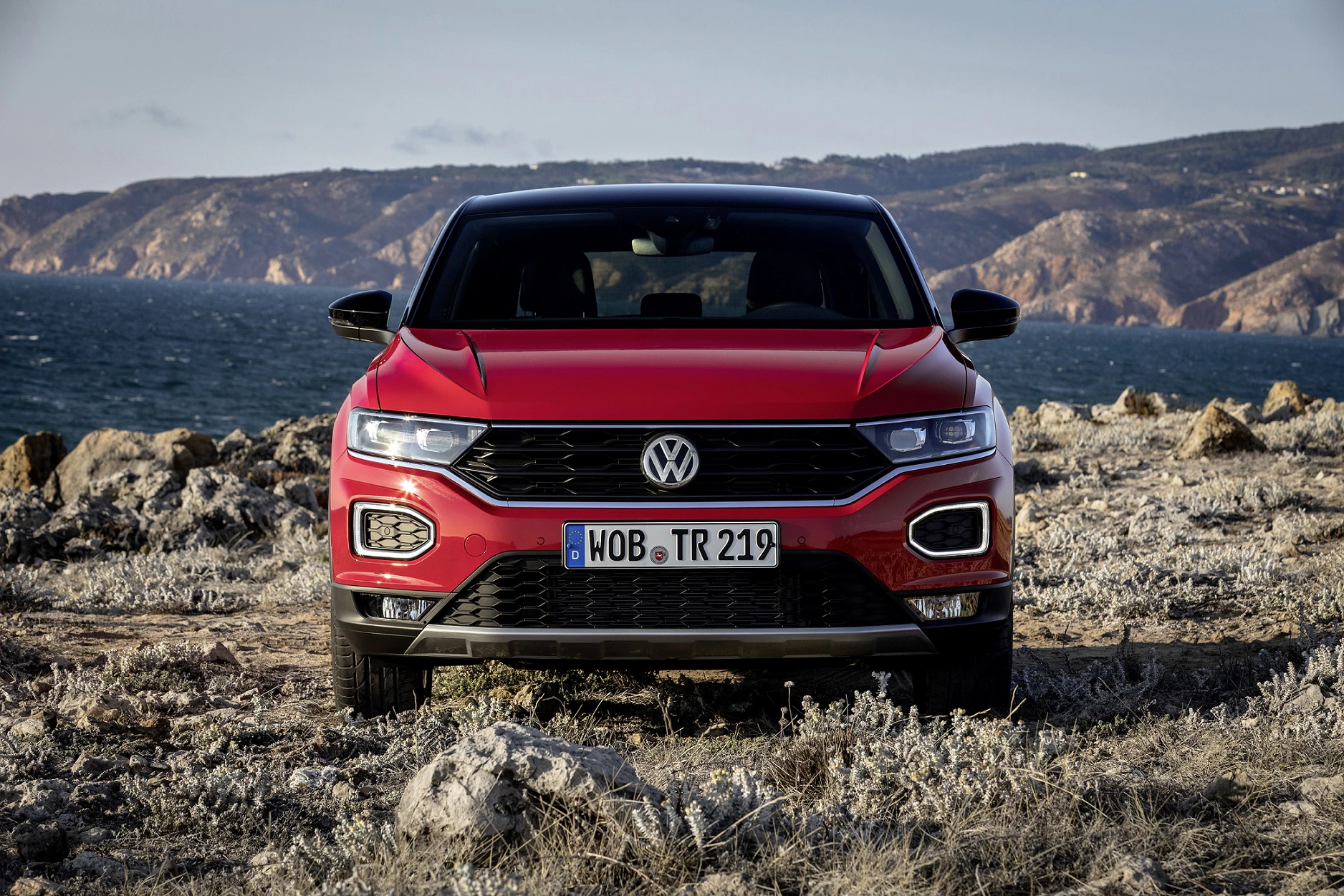 Roter Volkswagen T-Roc steht auf felsigem Gelände mit Meer und Bergen im Hintergrund, aufgenommen bei Tageslicht.
