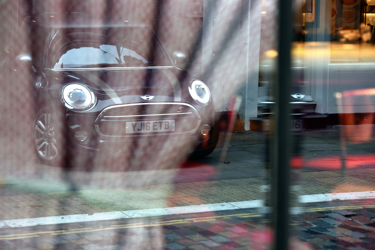 Ein Mini Cooper spiegelt sich in einem Schaufenster wider, umgeben von roten und gelben Lichtern auf einer gepflasterten Straße.