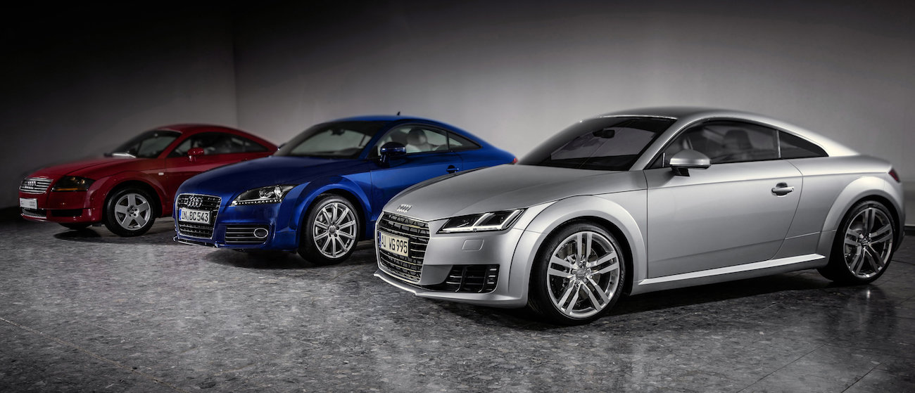 Drei Audi TT-Modelle in Rot, Blau und Silber stehen in einem Showroom nebeneinander auf grauem Boden.