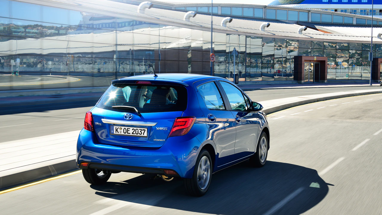 Blauer Toyota Kleinwagen fährt bei sonnigem Wetter auf einer Straße vor einem modernen Gebäude mit Glasfassade.