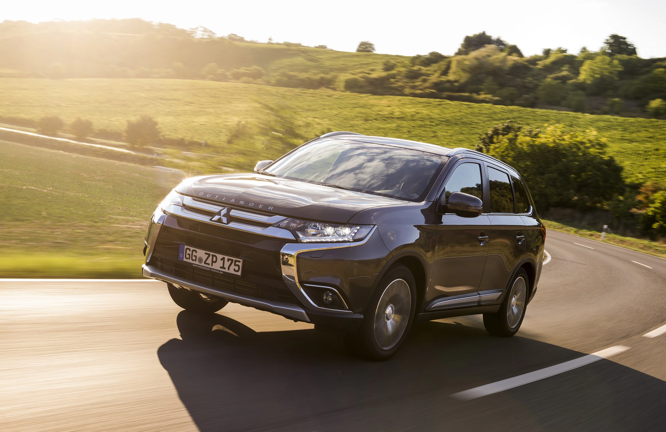 Dunkelgrauer SUV fährt auf einer Landstraße durch grüne Hügel und Weinberge bei Sonnenuntergang.