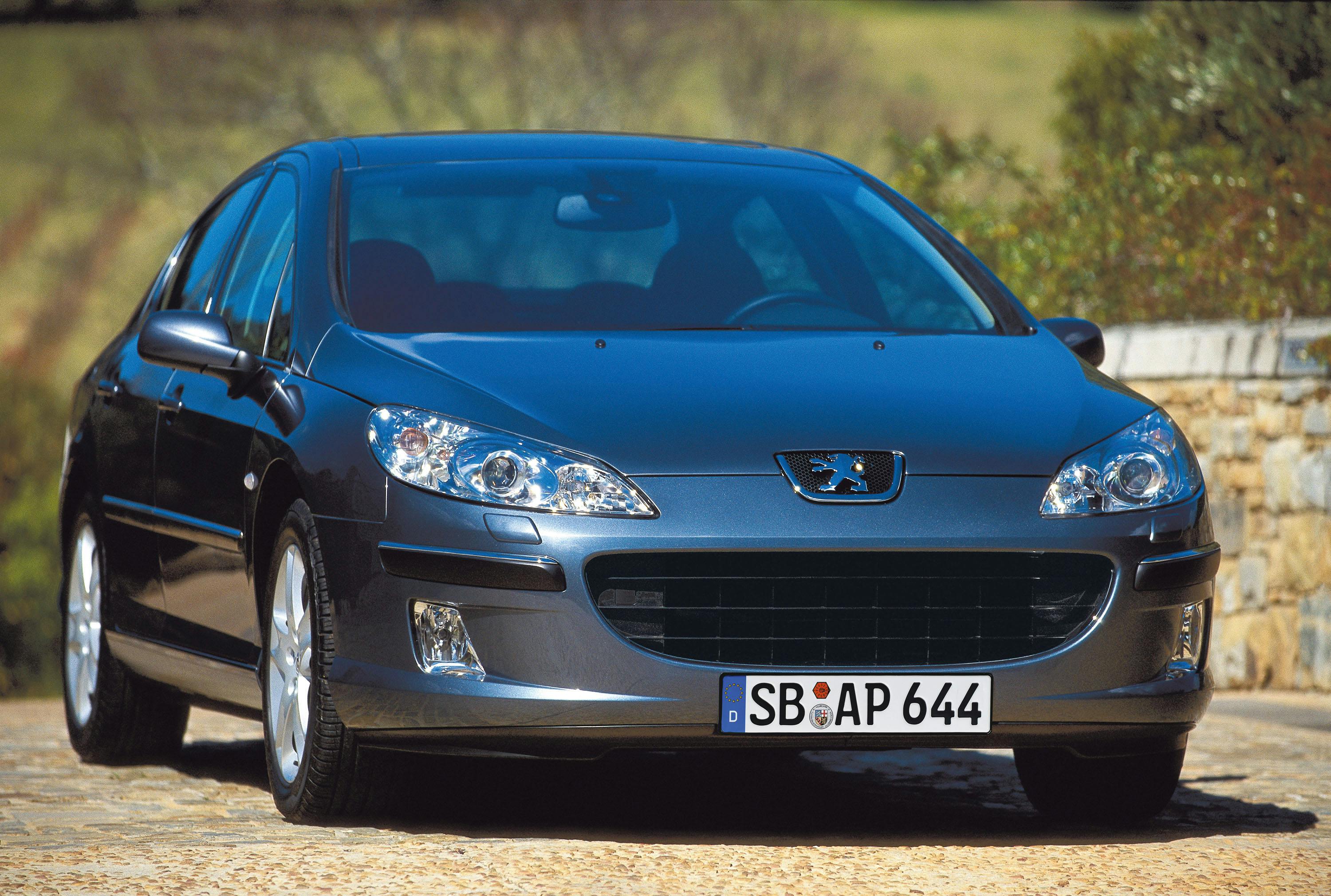 Dunkelgrauer Peugeot 407 mit deutschem Kennzeichen, geparkt auf gepflasterter Fläche vor einer Steinmauer und Bäumen.