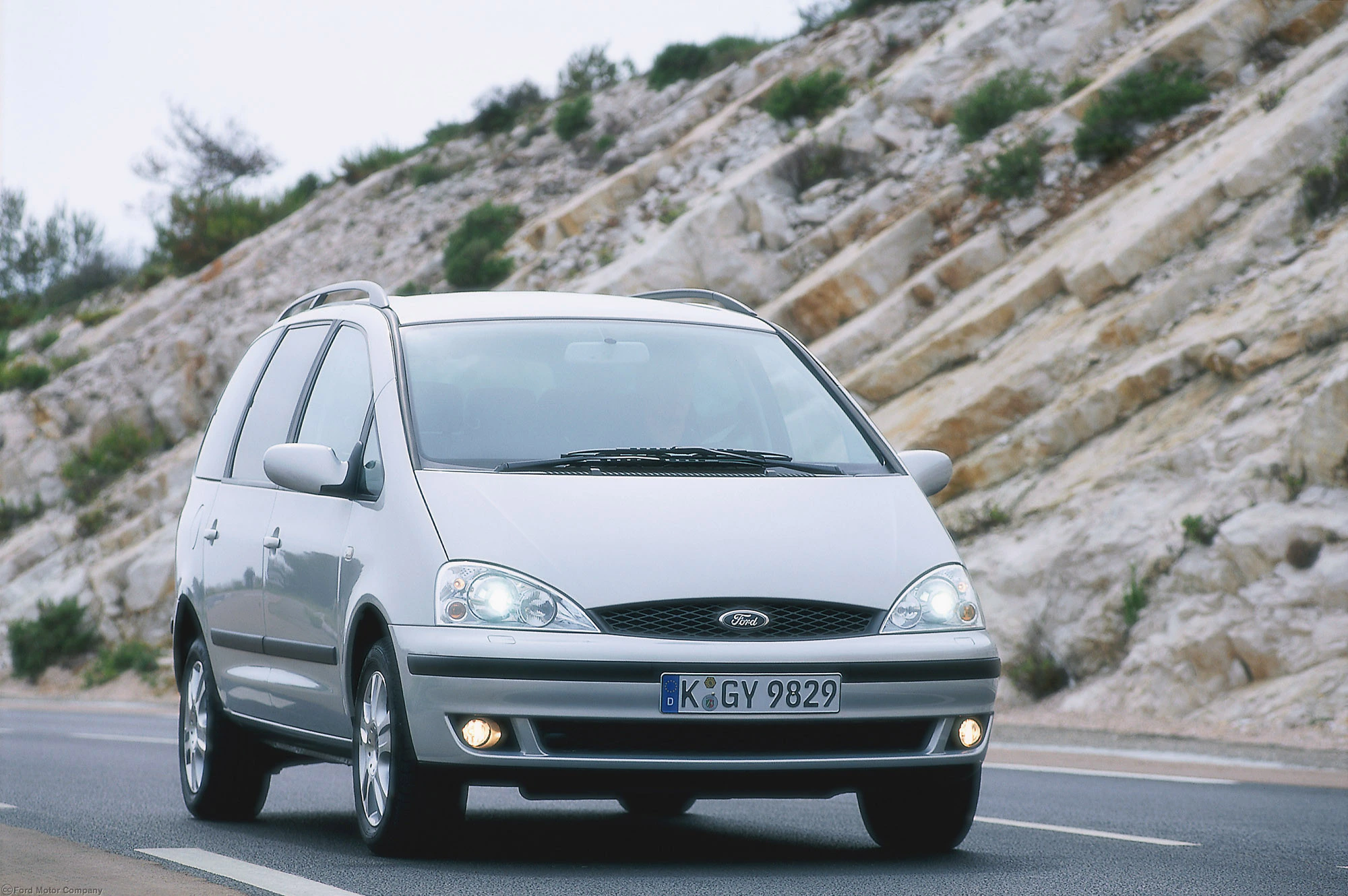 Silberner Ford Galaxy fährt auf einer Landstraße entlang felsiger Landschaft. Vorderansicht zeigt Scheinwerfer und Kennzeichen.