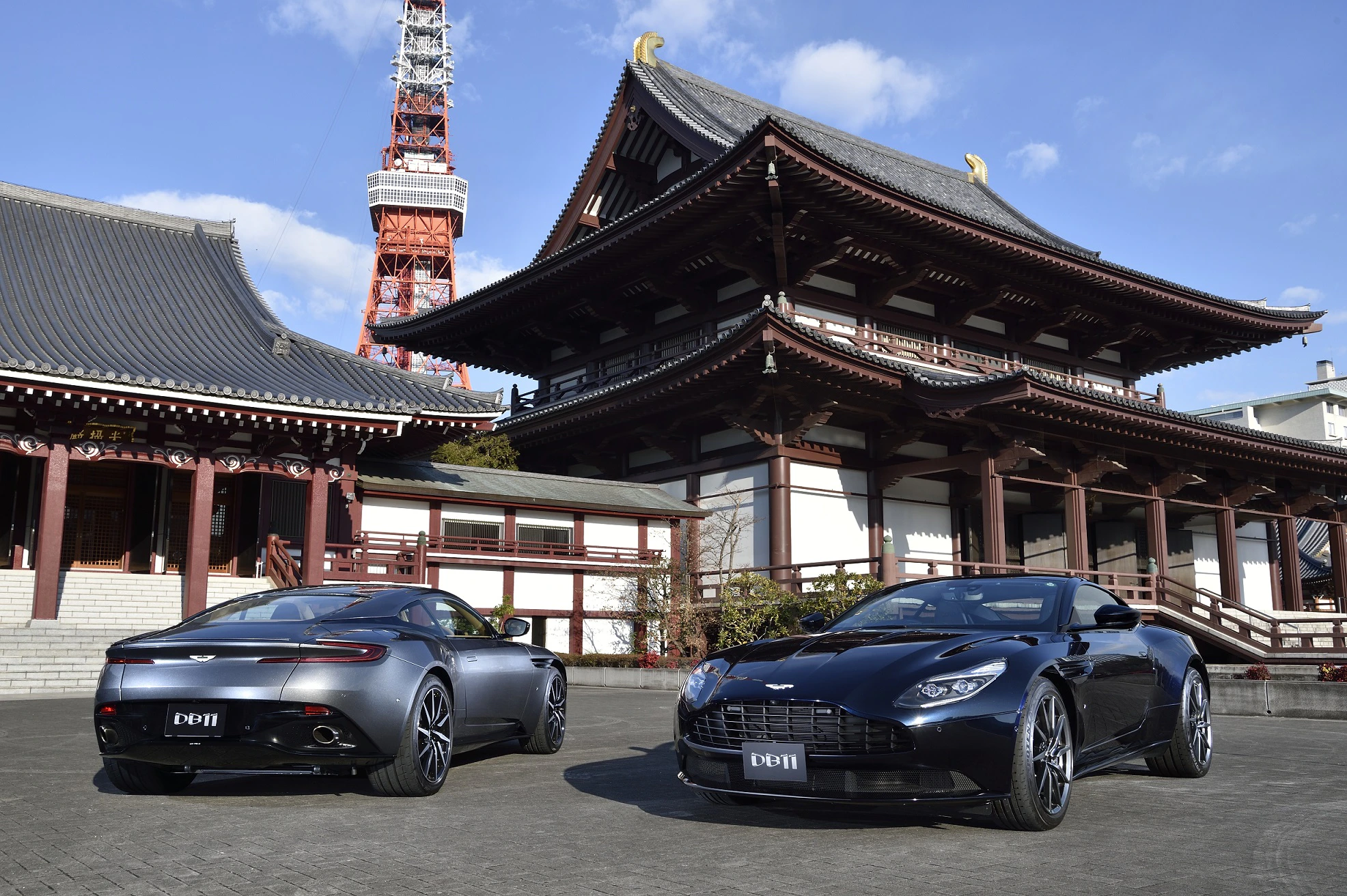 Zwei Aston Martin DB11 in Schwarz stehen vor traditioneller japanischer Architektur mit einem Turm im Hintergrund bei sonnigem Wetter.